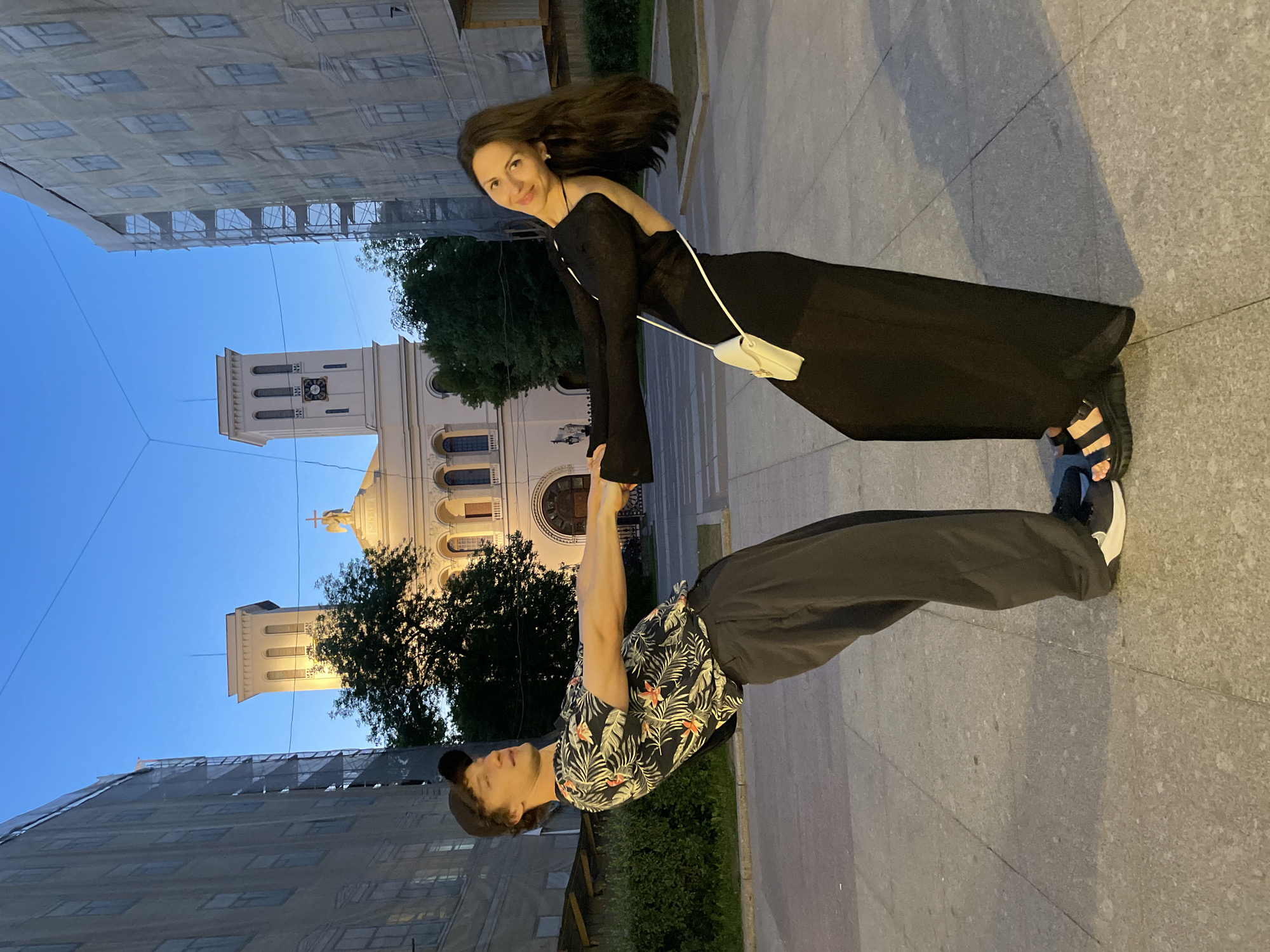 Two individuals, a male and a female, are depicted in a playful, leaning pose, holding hands on a paved surface. The setting is identified as Saint Petersburg, Russia, during twilight or late evening hours.
The male, positioned on the left, wears a black baseball cap, a black short-sleeved shirt with a tropical floral pattern (orange flowers and green foliage), loose-fitting black pants, and dark sneakers with white soles. He leans significantly backward, holding the female's left hand with his right, and looks slightly upward and to his right.
The female, on the right, has long dark hair and wears a long black dress with an open back and long sleeves, black platform sandals, and carries a small white crossbody bag with a gold clasp. She also leans backward, holding the male's right hand with her left, and smiles faintly while looking towards the viewer.
In the background, a large neoclassical building, confirmed as the Church of the Holy Myrrh-Bearers on Sredny Prospekt, Vasilyevsky Island, Saint Petersburg, is prominently visible. It features a central dome topped with a cross and two symmetrical bell towers, one of which displays a clock face. The building is light-colored and partially illuminated. Flanking the church are multi-story structures, both extensively covered in scaffolding and a light-colored protective mesh, indicating ongoing construction or renovation. The sky above is a clear, deep blue. The ground beneath the individuals is composed of grey paving slabs. Overhead utility wires are visible against the sky.