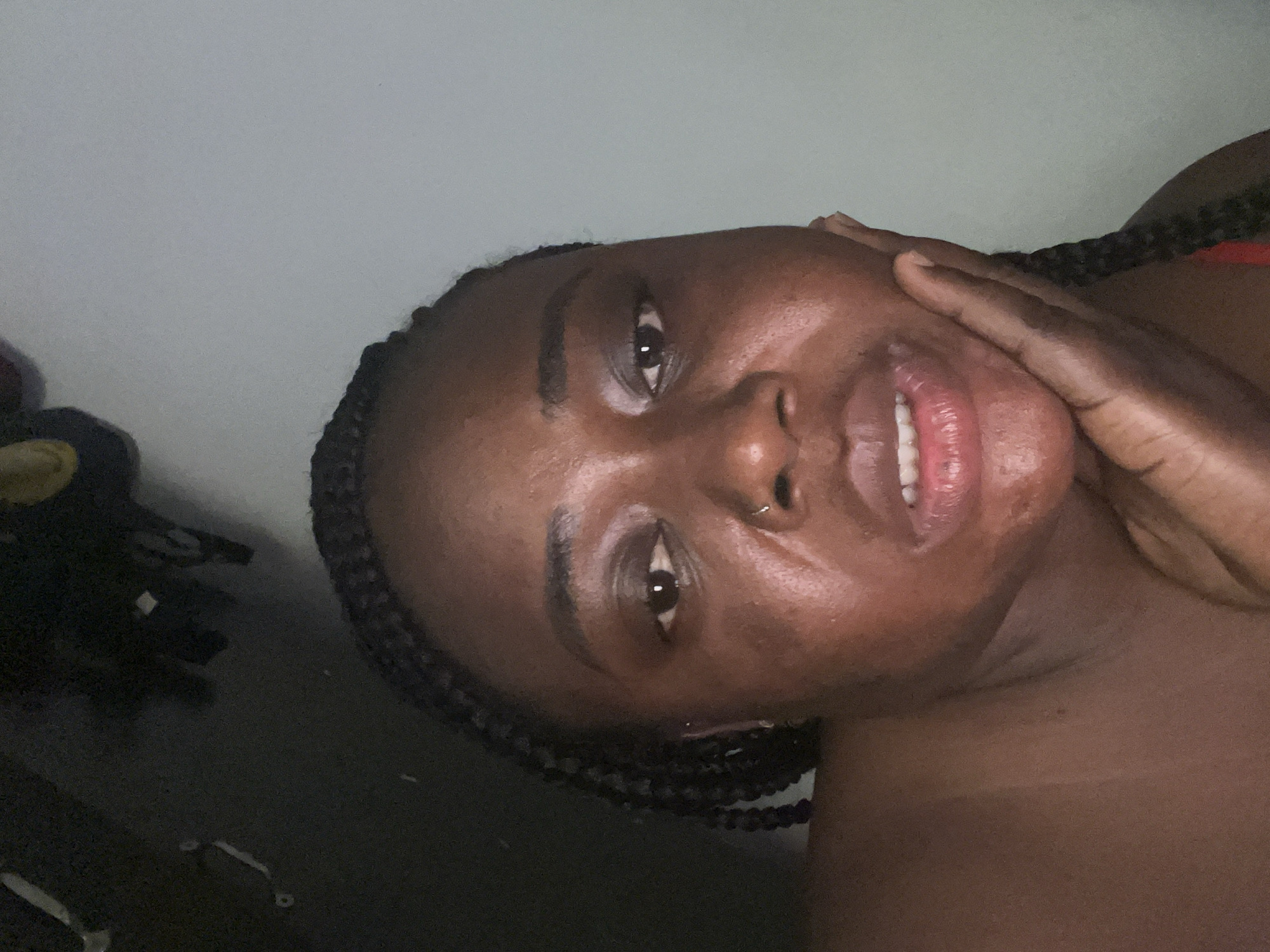 A close-up portrait shows a Black woman, likely in her 20s or 30s, looking directly at the camera with a soft, relaxed smile. Her lips are slightly parted, revealing her teeth. She has dark, braided hair framing her face and a small silver nose piercing visible on her right nostril. Her right hand is casually placed against her cheek and jawline. A hint of red clothing is visible on her right shoulder.
The setting appears to be indoors, with a plain, light grey or off-white wall forming the background in the upper portion of the image. The lower left corner shows a darker, indistinct area, possibly furniture or a dark surface. The lighting on her face is soft and even, suggesting an indoor light source. The image is slightly rotated, indicating a casual selfie. The woman's expression conveys a sense of calm and contentment. There are no specific visual cues within the image to identify the location as Abuja, Nigeria, beyond the context provided. No text is visible.