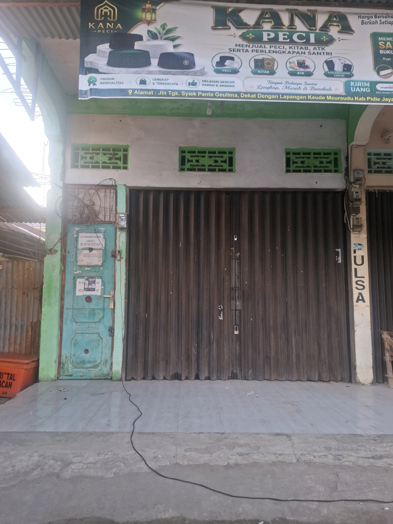 The image shows the storefront of a business named "KANA PECI" in Mesjid Tuha, Indonesia. The storefront features a large, dark brown roll-up door that is closed. Above the door, a banner displays the business name, along with images of various "peci" (traditional caps), books, and other items. Text on the banner indicates that the business sells "Peci, Kitab, ATK serta Perlengkapan Santri" (Peci, books, stationery, and student supplies) and emphasizes being a "Tempat Belanja Santri Lengkap, Murah & Barokah" (Complete, affordable, and blessed student shopping place). It also lists the address as "Jln Tgk. Syiek Pante Geulima, Dekat Dengan Lapangan Keude Meureudu Kab Pidie Jaya". To the left of the roll-up door is a teal-colored metal door with several notices attached, including one with the text "DI SEWAKAN KAMAR SANTRI" (Room for rent for students). An orange plastic container is visible to the far left of the image, in front of a corrugated metal fence. A black electrical cord runs from the door area across the tiled ground. The overall environment appears to be an outdoor street scene in a commercial or residential area.