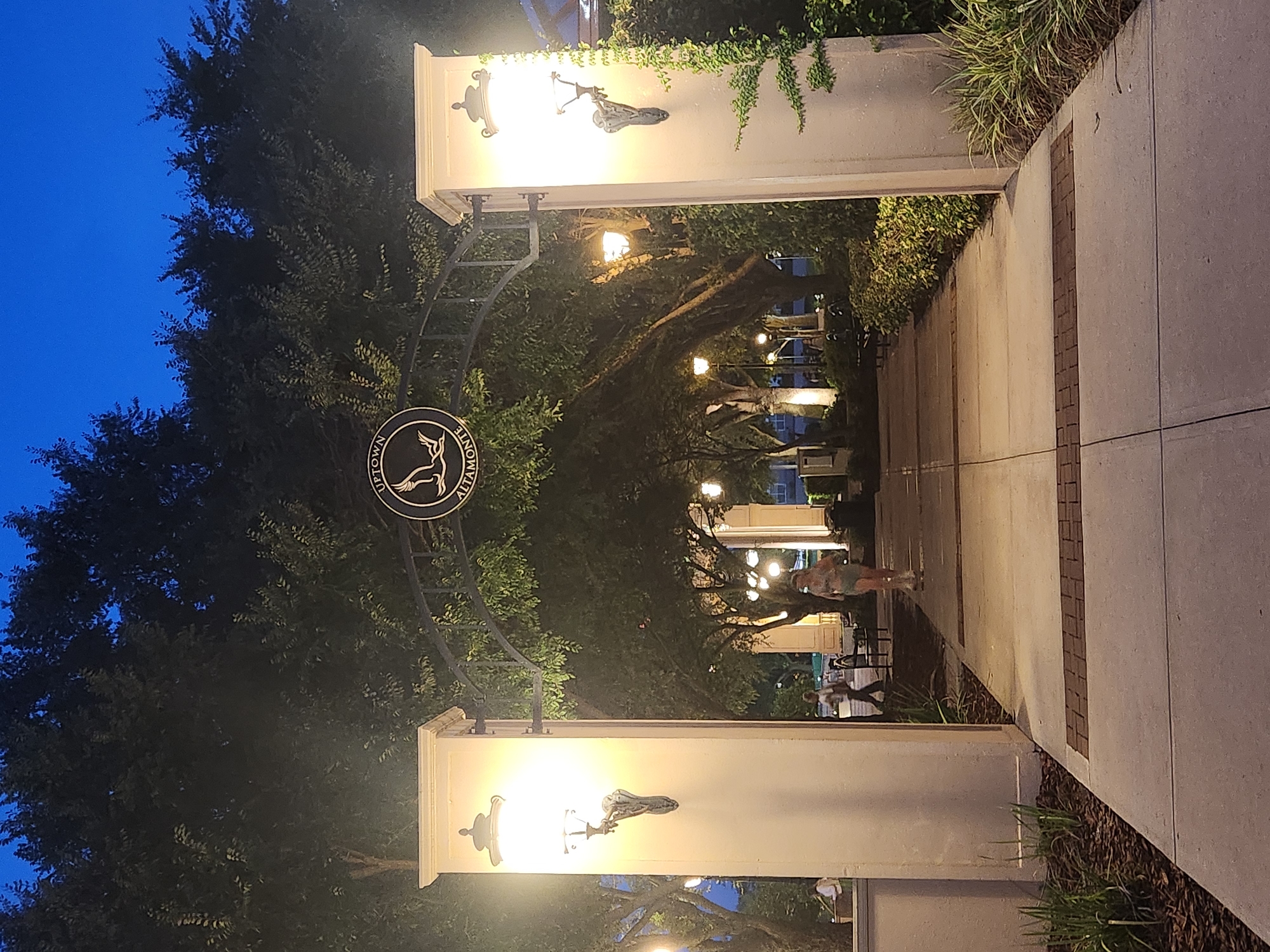 An arched entryway with a circular logo featuring a bird in flight, reading "UPTOWN ALTAMONTE," marks the entrance to a pathway in Altamonte Springs, Florida, United States of America. The scene is illuminated by ornate wall sconces on flanking pillars and further down the path by lampposts, creating a warm glow against the twilight sky. Trees with dense foliage line the pathway, casting shadows and contributing to the ambiance. A woman in athletic wear is seen walking away from the camera on the right side of the path, while another person is partially visible in the background on the left. Benches are placed along the walkway, suggesting a public space designed for leisure. The pathway itself is paved with concrete, interspersed with decorative brick accents.