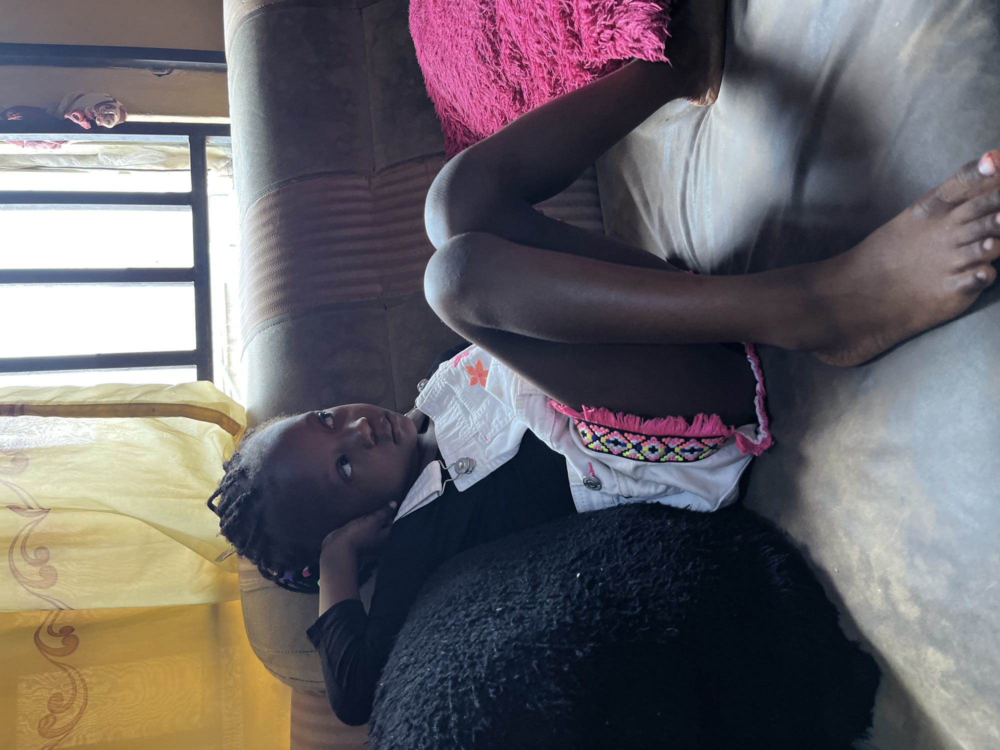 A young person lies on a tan couch with a pink pillow. The individual is wearing a black top and a white jacket with pink trim. The person's bare legs are crossed. A black, furry object is near the individual. Behind the couch is a window with yellow curtains. The location is Madalla, Nigeria.