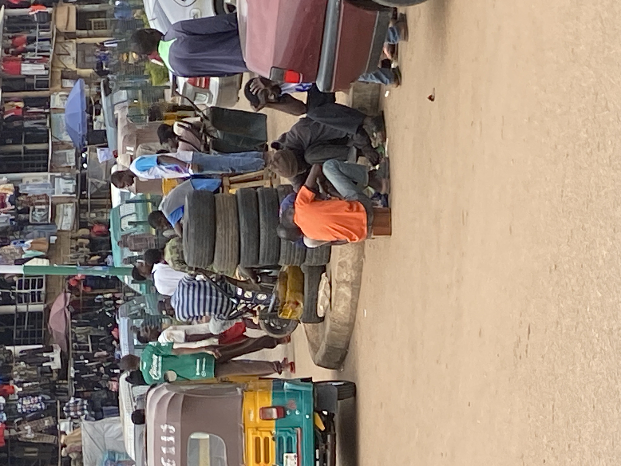This rotated outdoor image depicts a bustling street market scene in what appears to be Bukuru, Nigeria. The setting features unpaved ground and multi-story buildings lining the left side, with various goods displayed on shelves, suggesting an active commercial area.
Numerous people, predominantly men, are visible throughout the frame. Some individuals are standing, while others are seated on the ground or low structures, engaged in daily market activities. In the mid-ground, a man in an orange t-shirt sits beside a large stack of vehicle tires, indicating a tire sales or repair stall. Another person in a blue and white striped shirt is partially visible nearby. Farther down, a man wearing a green t-shirt is seen near a brown and yellow tricycle.
Various objects populate the scene, including several vehicles. A red car is parked on the right, and a lighter-colored car is visible in the upper right. A prominent brown and yellow tricycle (a common auto-rickshaw or Keke Napep in Nigeria), with the number "5115" faintly visible on its side, is in the lower left foreground, attached to a smaller blue-green vehicle. A blue open umbrella provides shade on the left side of the market. The presence of multiple tires, vehicles, and a variety of goods suggests a busy trading environment.
The scene takes place during the daytime, under clear skies, with natural light illuminating the area. The general atmosphere is that of typical market commerce, with people going about their routines.