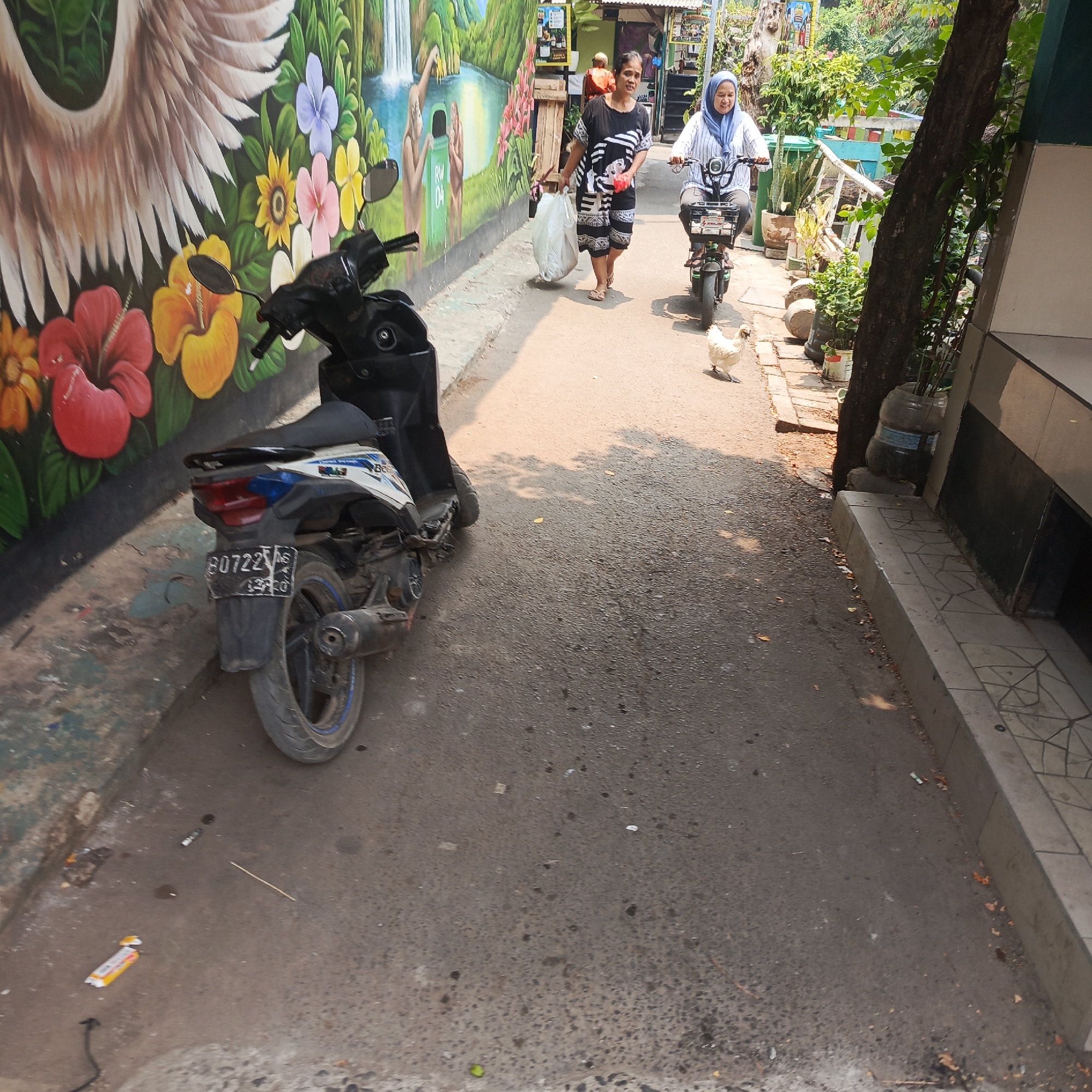 A paved alleyway in the residential area of Tebet, South Jakarta, Indonesia is depicted. The alley is bordered by a building with a tiled sidewalk on the right and a vibrantly painted mural on the left. The mural features large, colorful flowers and a depiction of a waterfall with figures. A black motorcycle is parked on the left side of the alley, facing away from the camera. In the center of the alley, a woman wearing a patterned dress walks towards the camera, carrying a large white plastic bag. To her right and further down the alley, a woman wearing a hijab and a striped shirt rides a green electric scooter. A white chicken is walking on the alleyway in front of the scooter. The ground is littered with small debris, including a discarded cigarette lighter and a piece of yellow packaging. The scene is illuminated by sunlight, casting shadows on the alleyway.
