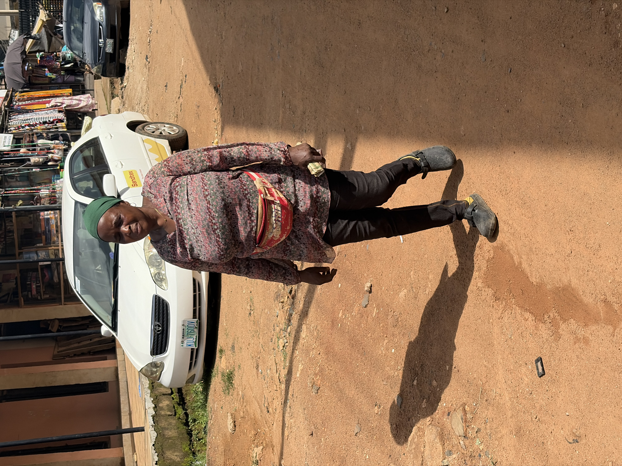 The image captures a street scene in Jos, Nigeria, on a sunny day. A woman, dressed in a colorful patterned shirt and black pants, is standing on a dirt road. She wears a green headscarf and a red fanny pack around her waist. She holds a wad of cash in her right hand. To her left, a white car is parked, with the text "Sandtex" visible on its side. Behind the car, market stalls with various goods are set up, including clothing and an umbrella. The overall impression is of a bustling marketplace. The lighting suggests it is daytime, and the shadows indicate a bright, sunny environment. The woman's expression appears neutral. The license plate on the car is visible and reads "KUJ-760DV".