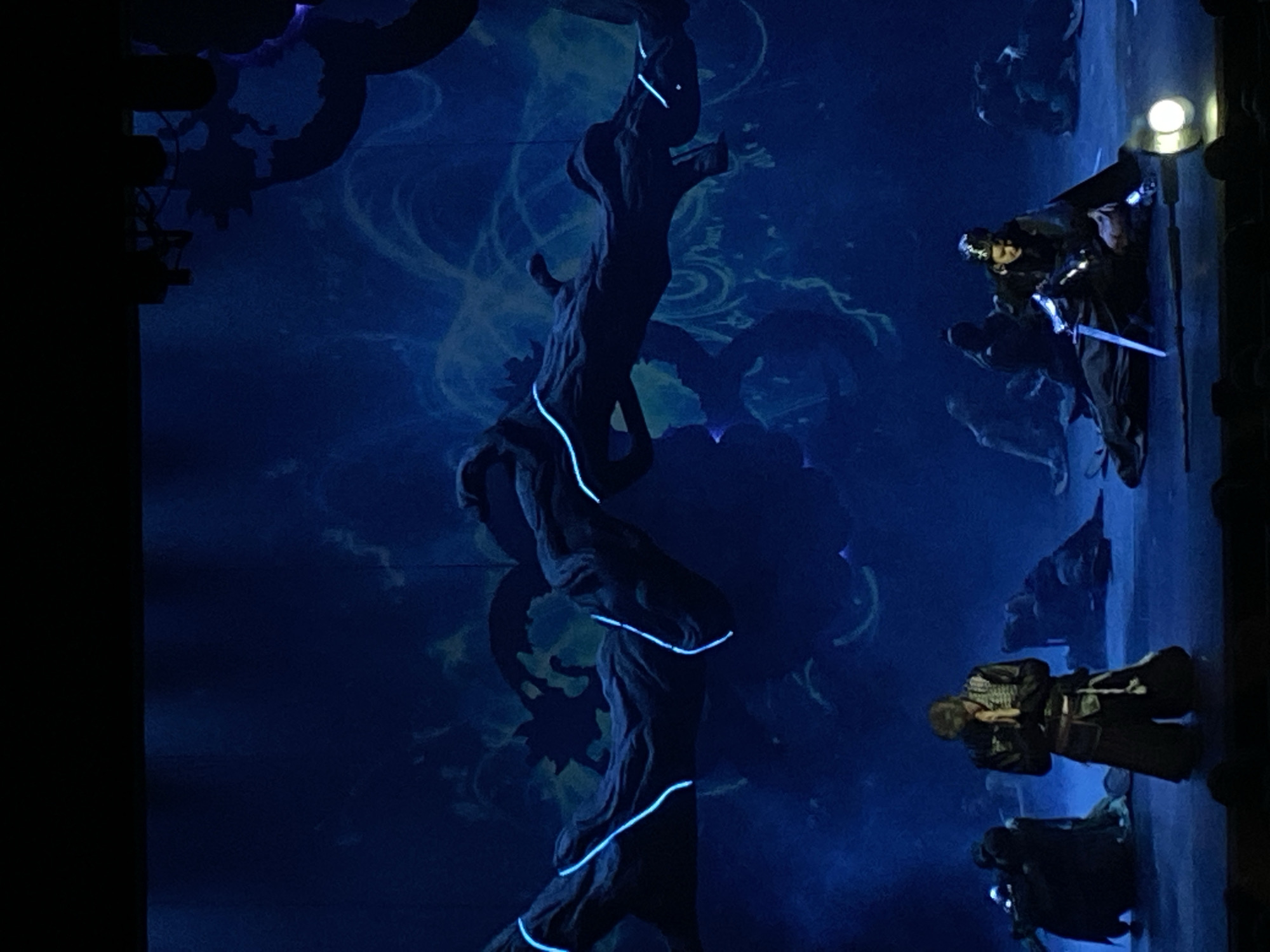 The image depicts a stage performance with a dark blue color scheme. A gnarled tree trunk with illuminated blue accents stands prominently. Two figures dressed in what appears to be Japanese samurai attire are present on the right side of the frame. The location is likely a theater in Заволжье, Russia, inferred from the prompt.