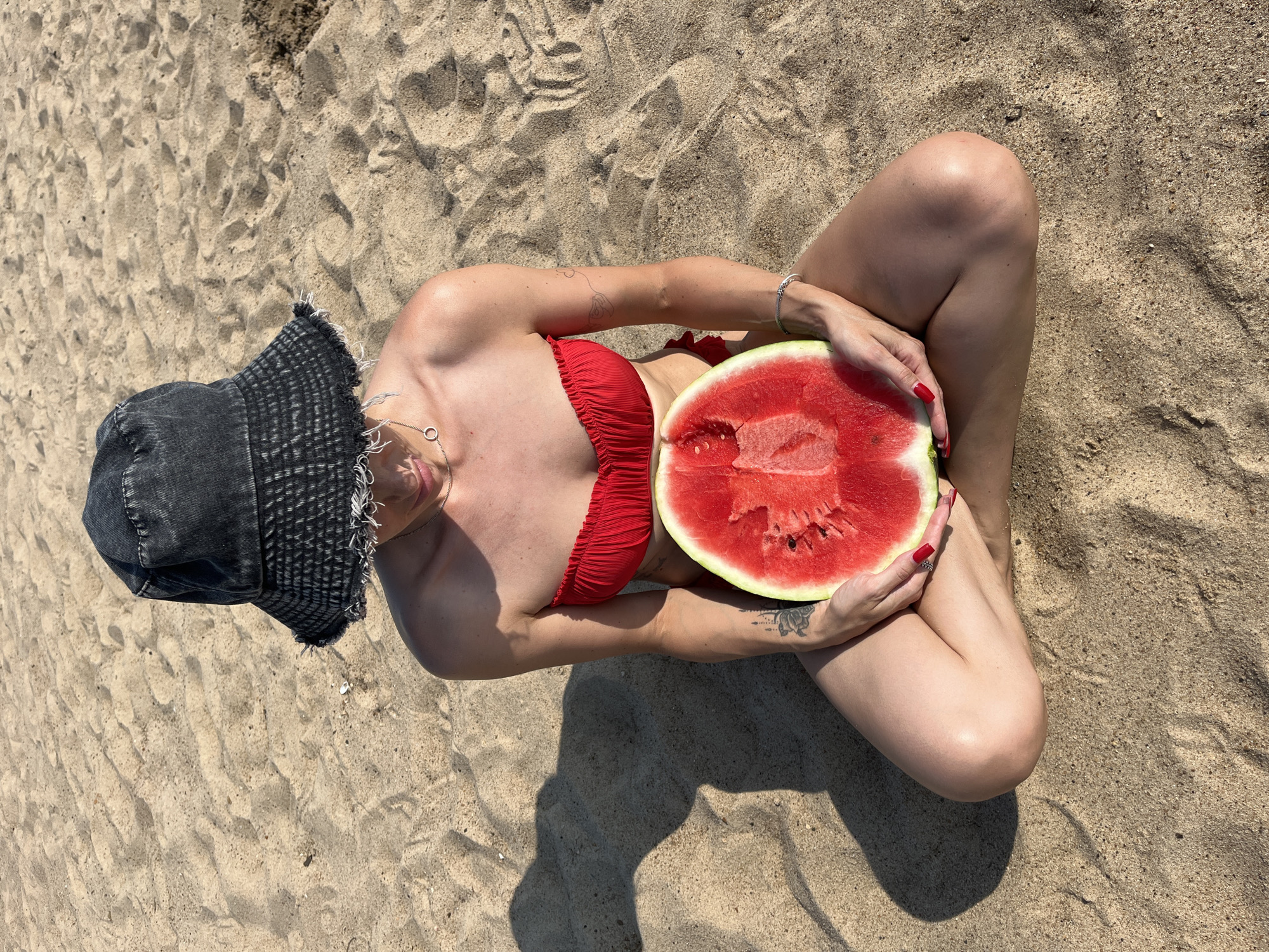A person sits cross-legged on a sandy beach, holding a large slice of watermelon. They are wearing a red strapless bikini top and a dark denim bucket hat. The person is looking down at the watermelon. The environment is a beach with light brown sand.