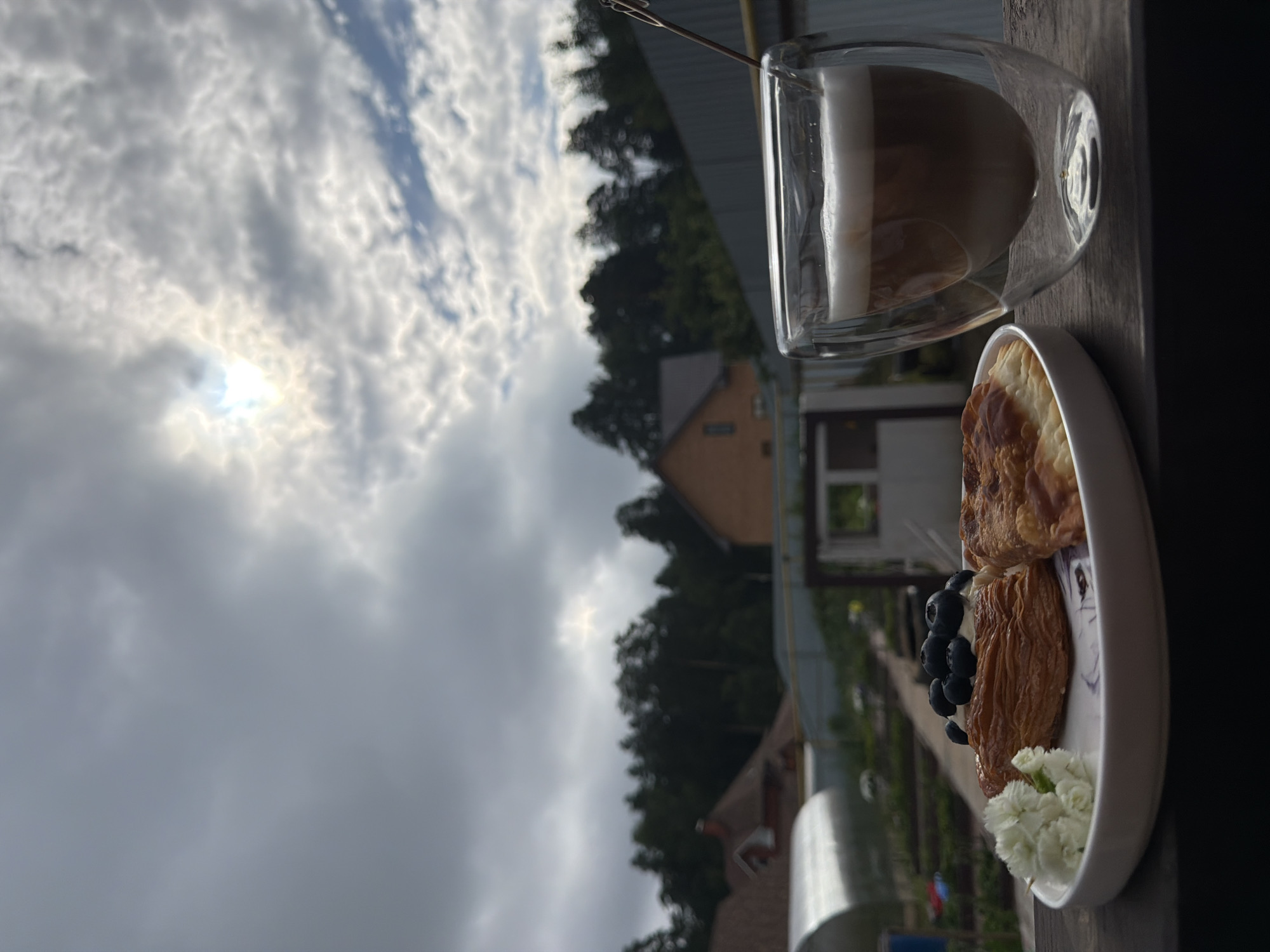 A plate with two pastries and a glass of coffee sits on a wooden surface. The pastry on the left is topped with blueberries and cream, while the one on the right is golden brown and flaky. The coffee has layers of milk and espresso. In the background, a suburban landscape is visible, with houses, trees, and a greenhouse under a cloudy sky. The sun is peeking through the clouds.