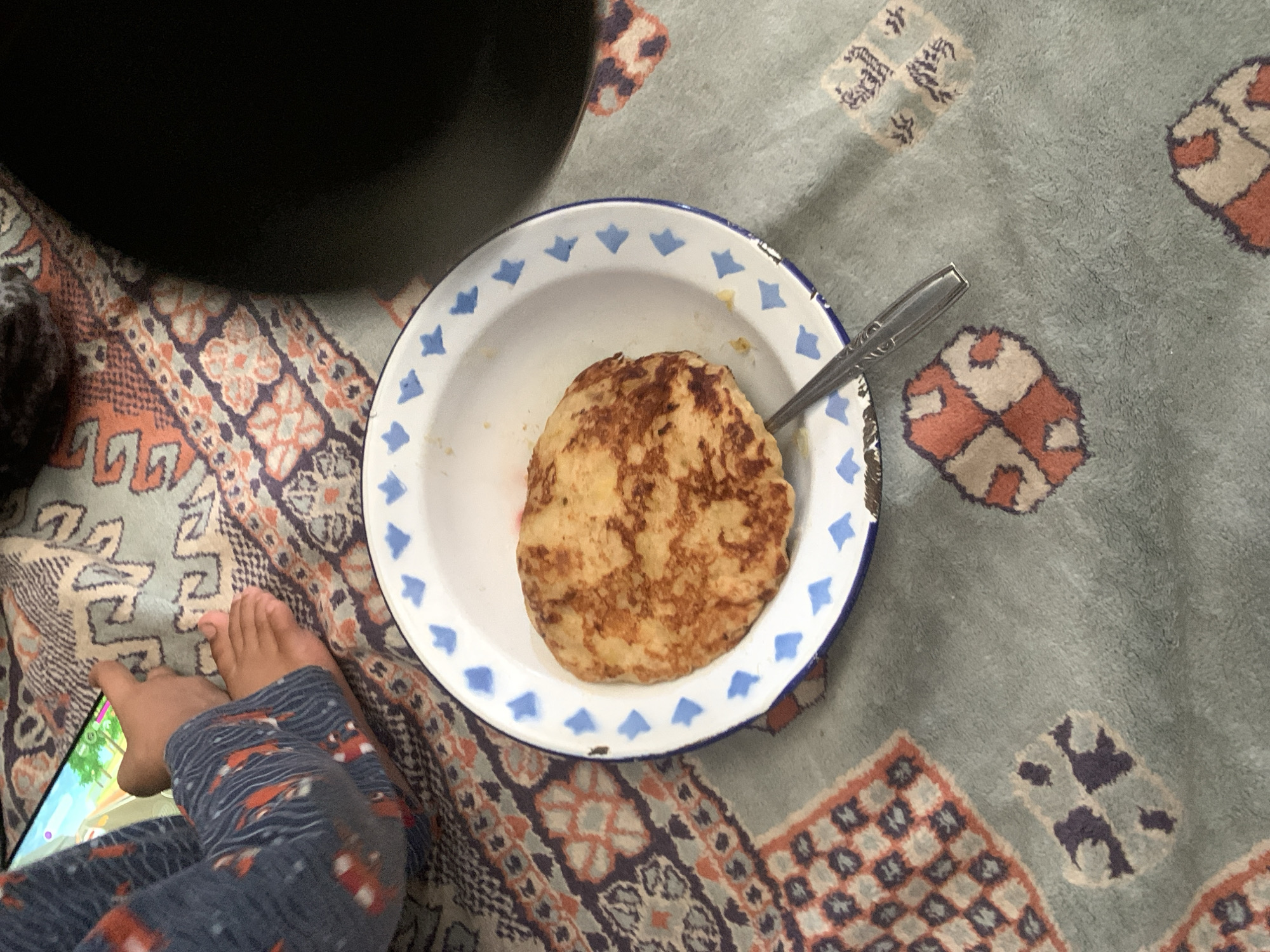 The image shows a close-up of a child's feet and legs wearing patterned pajamas, resting on a patterned blanket. A tablet is visible, displaying a colorful game. In the foreground, a round, golden-brown pancake sits in a white enamel bowl with blue trim. A silver fork rests on the pancake and extends out of the bowl. The blanket has a complex geometric pattern with shades of blue, orange, and tan. The scene appears to be indoors, with a dark, out-of-focus object in the upper right corner possibly being a piece of furniture or another person. There is no specific indication of the location being Sungai Meriam, Indonesia.