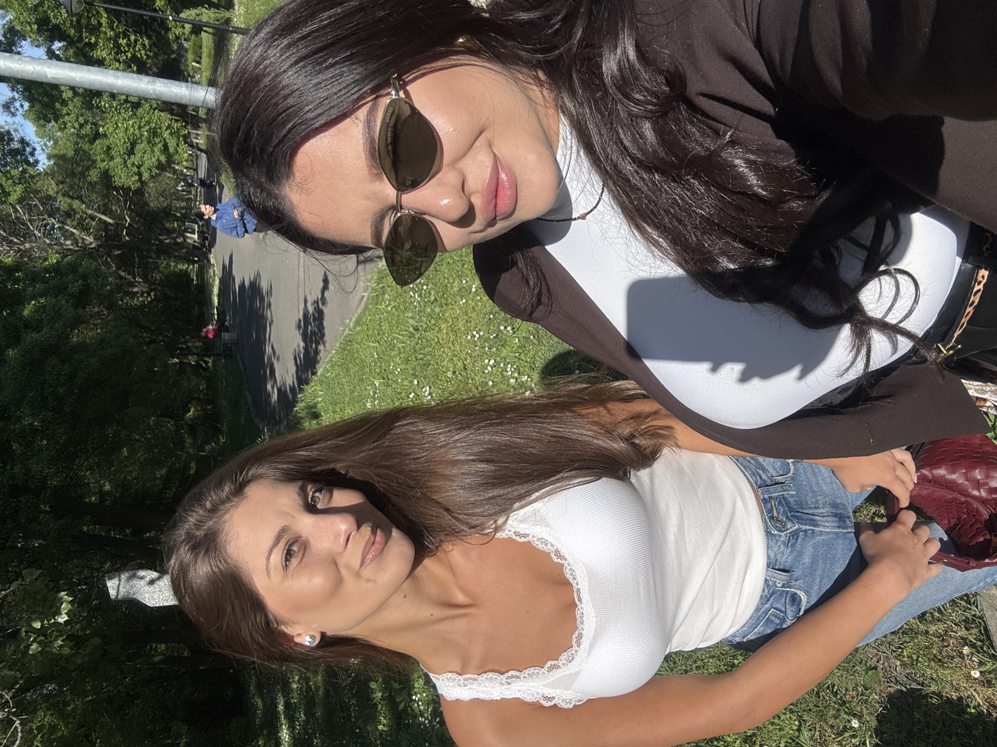 Two women are posing for a selfie outside in a park. The woman in the foreground is wearing a white tank top with lace and jeans. The woman in the background is wearing sunglasses, a white turtleneck, and a brown jacket. They are sitting on the grass in a park-like setting with trees and a paved walkway in the background. The location is inferred to be Burgas, Bulgaria, from additional context not provided in the image.