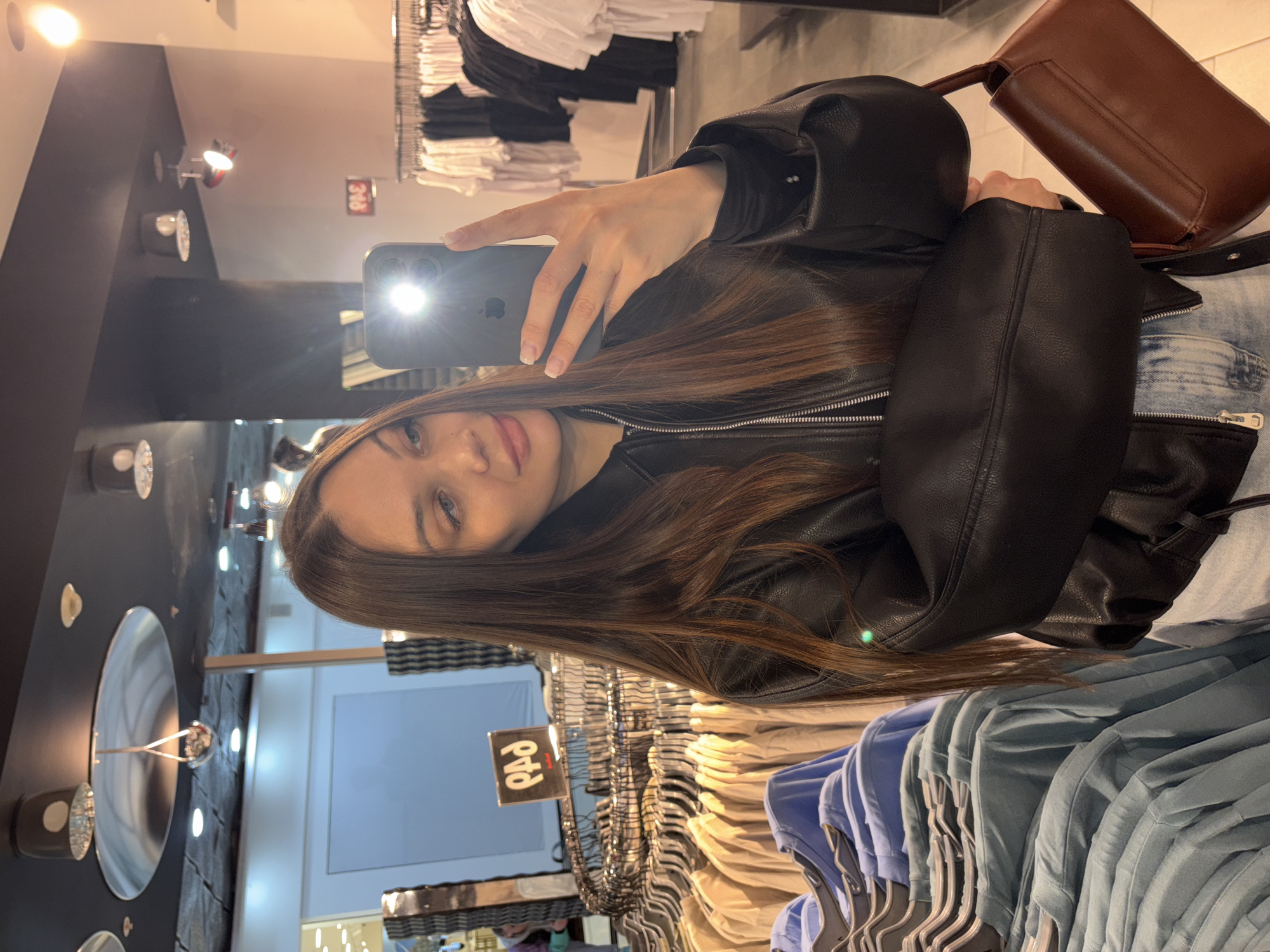 A young woman in a black leather jacket is taking a selfie in what appears to be a clothing store. She is holding a phone in her right hand, which is creating a bright glare on the lens. The background shows racks of clothing, suggesting she is shopping. Based on the reflection of the 'РАД' sign in the mirror, the location may be in Yekaterinburg, Russia. Her long brown hair is visible in the foreground. A brown purse is partially seen next to her. She is wearing a light-colored denim jacket or top underneath the leather jacket.
