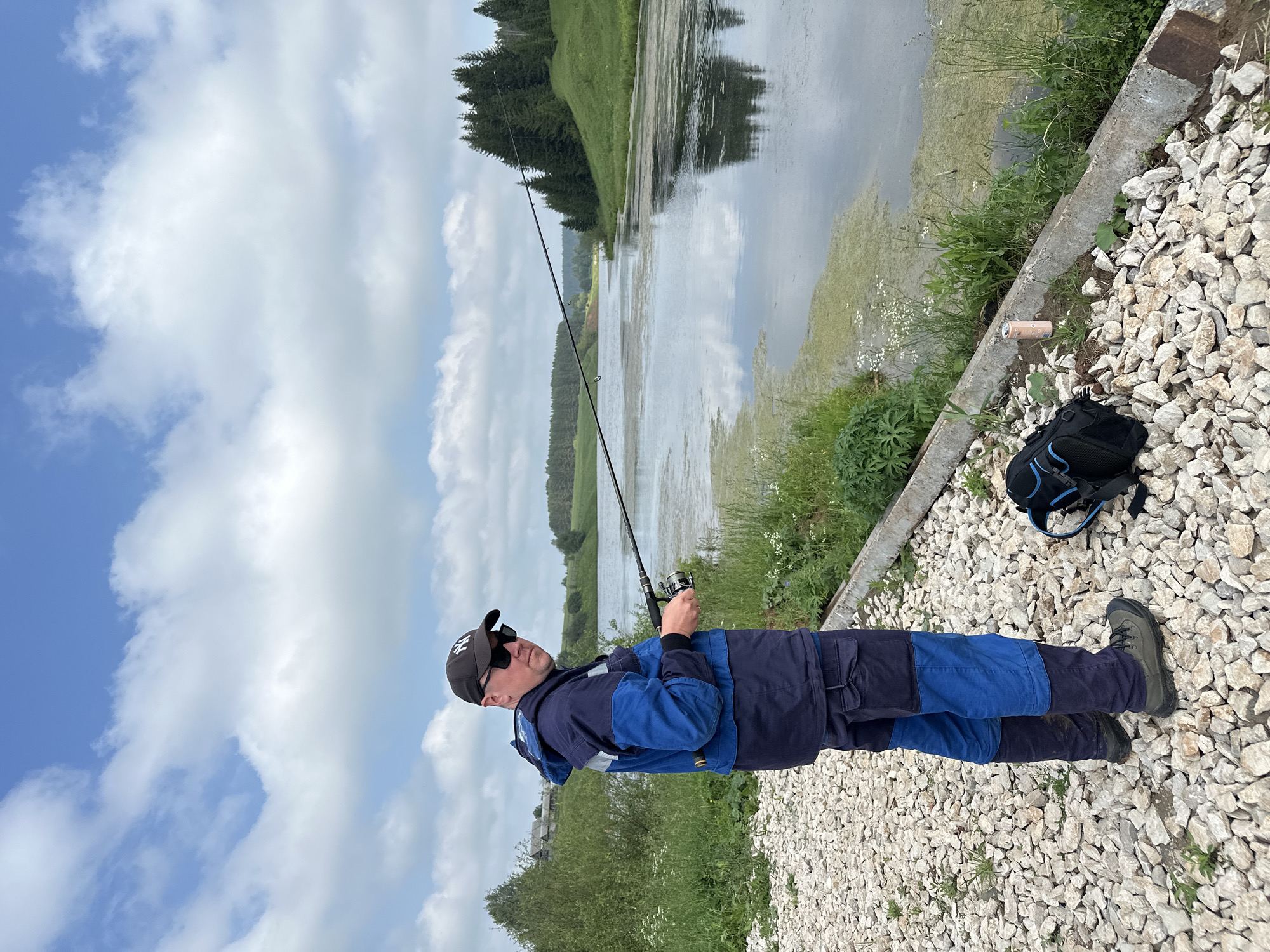 A man, wearing a dark baseball cap, sunglasses, a blue and dark blue jacket, and blue pants, stands on a gravel path, fishing in a calm body of water. His fishing rod is extended over the water, and he is holding the reel. A black backpack with blue accents lies on the gravel near his feet. The background features a gentle green slope with trees at the top, and the sky is filled with scattered white clouds. In the distance, there appears to be a small village. The scene suggests a tranquil outdoor setting, possibly in a rural area like the city of Sredneye Kechevo, Russia.