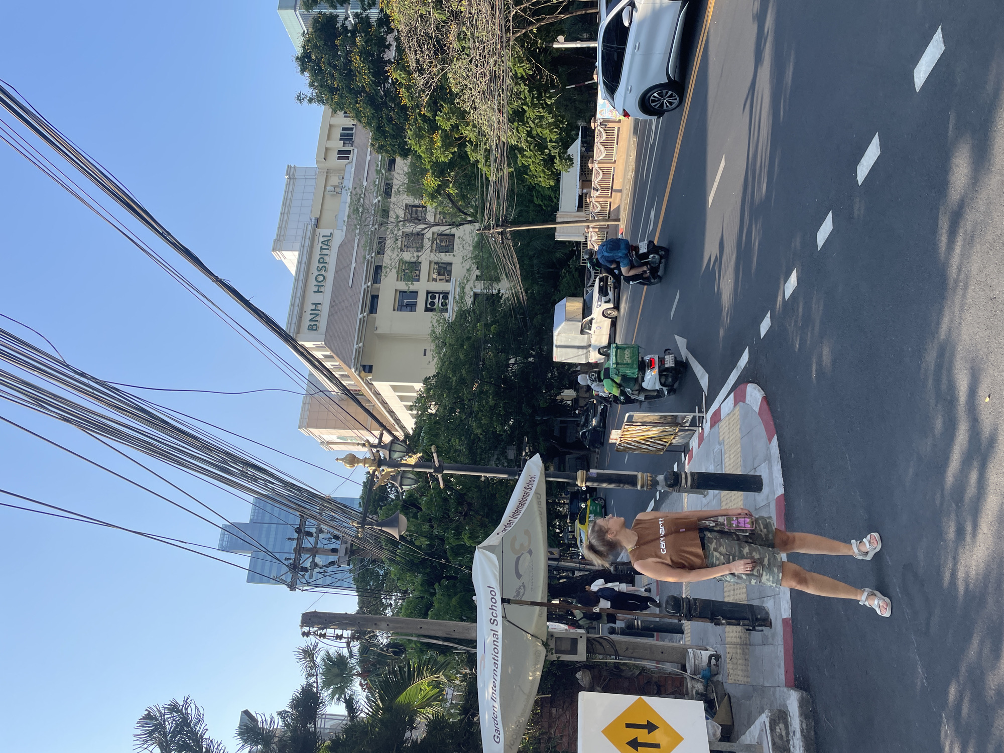 The image depicts a street scene in Bangkok, Thailand. The orientation of the image is sideways. In the foreground, a woman walks along the sidewalk wearing a brown Carhartt t-shirt, camouflage shorts, and white sandals. A street sign with yellow arrows pointing in opposite directions is visible. An umbrella with "Garden International School" printed on it stands nearby. Power lines are strung overhead.
In the background, a street with cars and motorbikes is seen. A white building labeled "BNH Hospital" is also in the background, along with other buildings and trees. The sky is clear and blue.