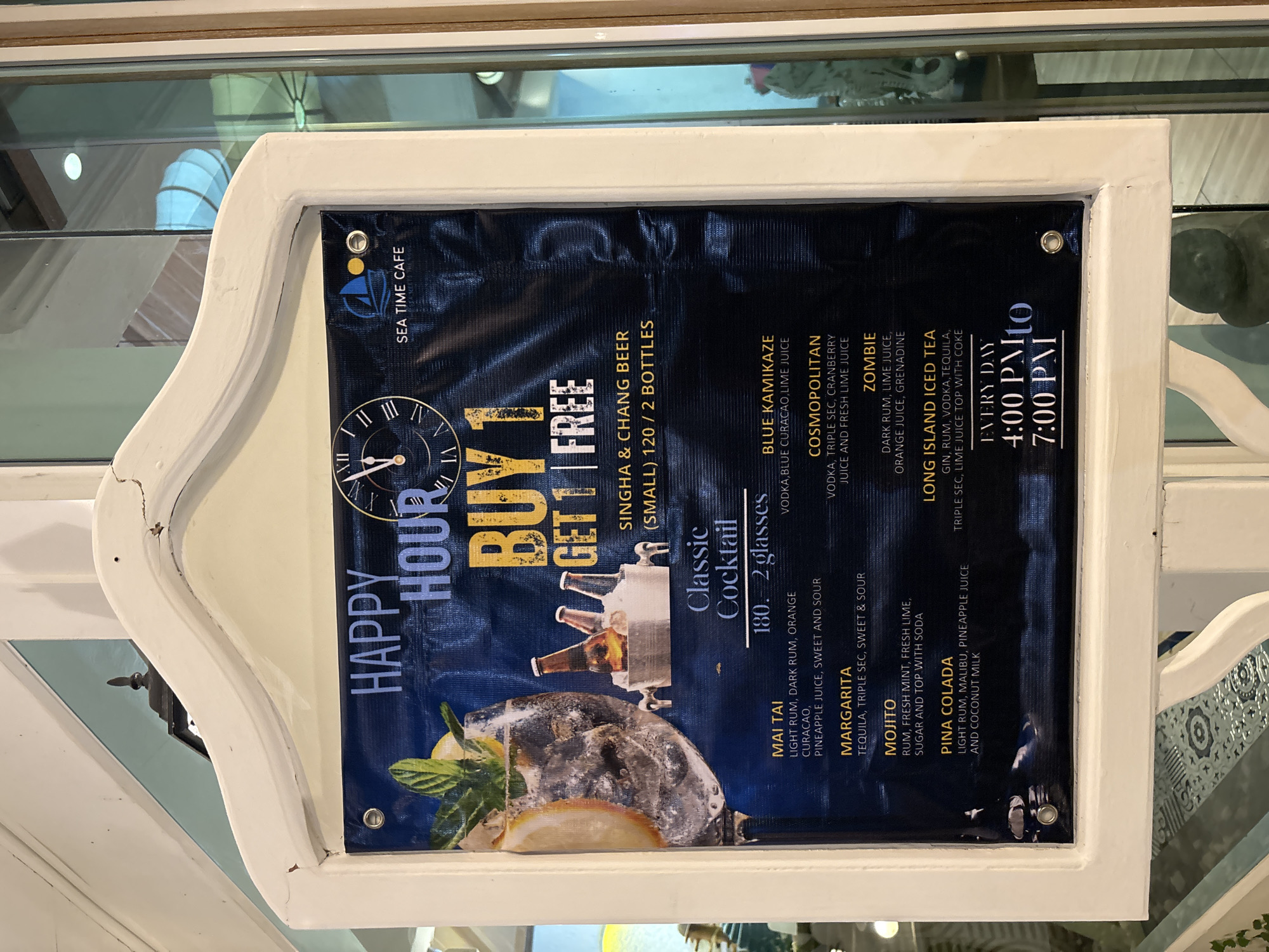 The image shows a close-up of a white wooden frame displaying a dark blue banner with a happy hour menu for "Sea Time Cafe" in Chalong, Thailand. The banner features text and graphics promoting a "BUY 1 GET 1 FREE" offer on Singha & Chang beer. It also lists various classic cocktails, including Mai Tai, Margarita, Mojito, Pina Colada, Blue Kamikaze, Cosmopolitan, Zombie, and Long Island Iced Tea, along with their ingredients and prices. The happy hour is advertised to be from 4:00 PM to 7:00 PM daily. A large image of a cocktail with mint and a lemon slice is prominently displayed on the left side of the banner, next to a small ice bucket with bottles of beer. The lighting suggests an indoor environment, possibly a cafe or bar.