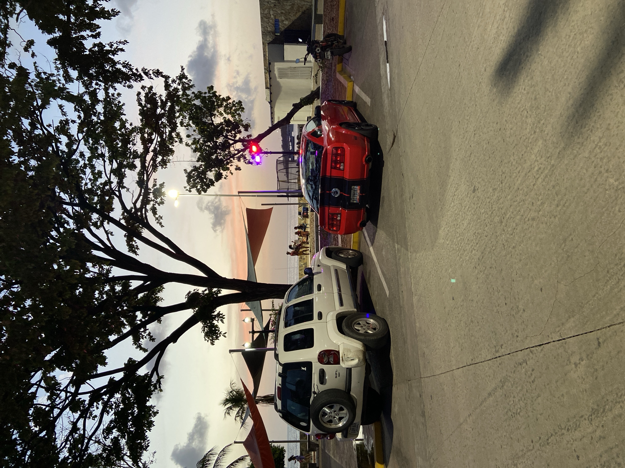 The image depicts a parking lot scene in Juan Griego, Venezuela, near the coast at dusk. In the foreground, a red Ford Mustang and a white Jeep Liberty are parked. The Mustang is facing away from the camera, showcasing its rear with black racing stripes. The Jeep, also facing away, has a spare tire mounted on its rear. Further back, the ocean is visible with a few people on the beach, some under colorful canopies. A street lamp with a flashing red and blue light is positioned behind the Mustang, casting a colored glow. A motorcycle is parked to the right of the Mustang. The sky is a mix of pastel colors, indicating sunset. Trees with dark foliage frame the upper portion of the image. The parking lot surface is paved concrete with visible cracks and tire marks.