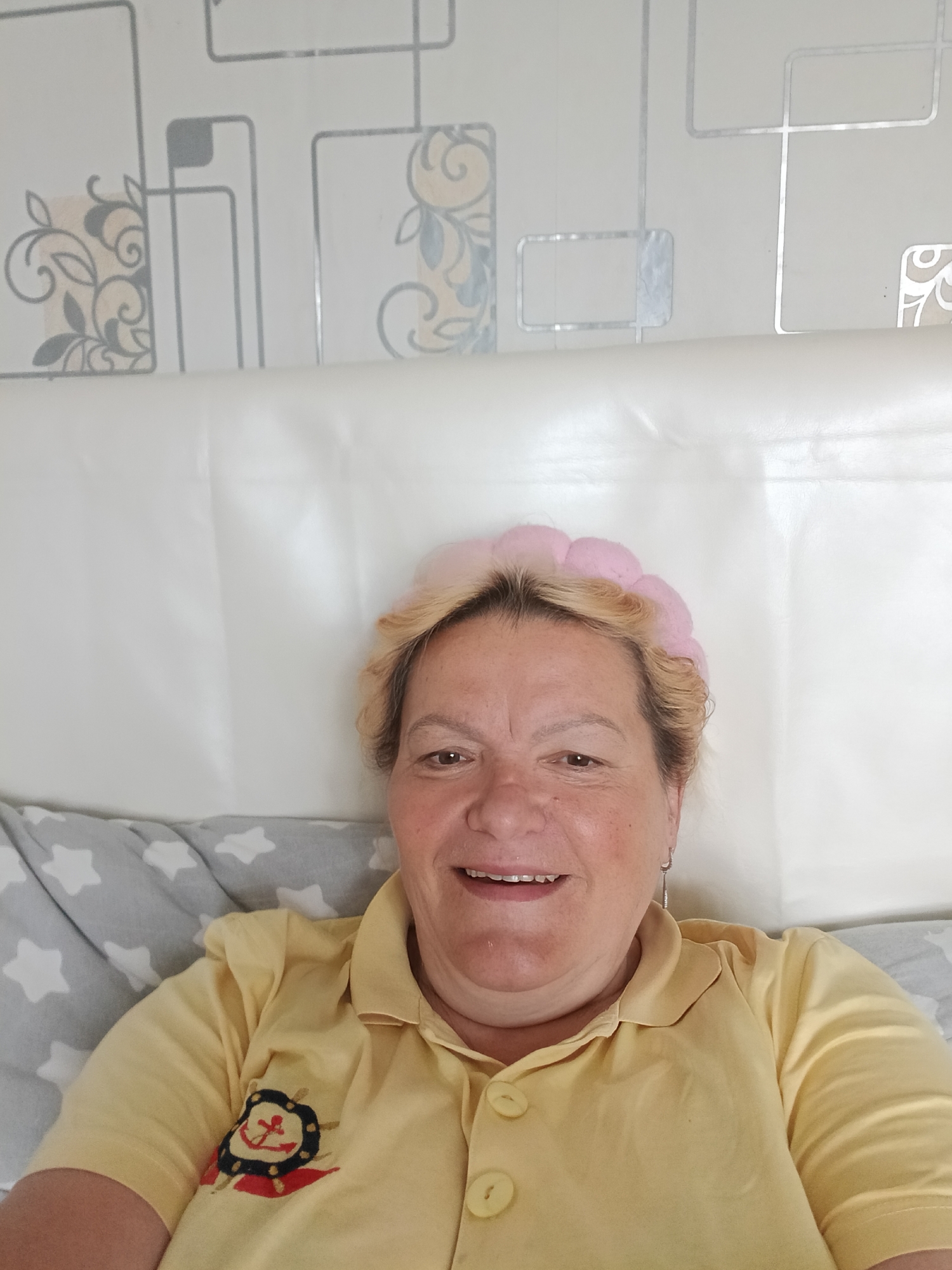 A woman is in bed, wearing a yellow collared shirt with an anchor emblem on the left side, and a pink headband. She is smiling at the camera. The bed is made with a pillow that has light gray stars on a medium-gray background. The headboard of the bed is a solid white, behind which is a wall with a geometric pattern of silver, light gray and white. The location is unknown, and there is no evidence to suggest the location is Красный Маяк, Russia.
