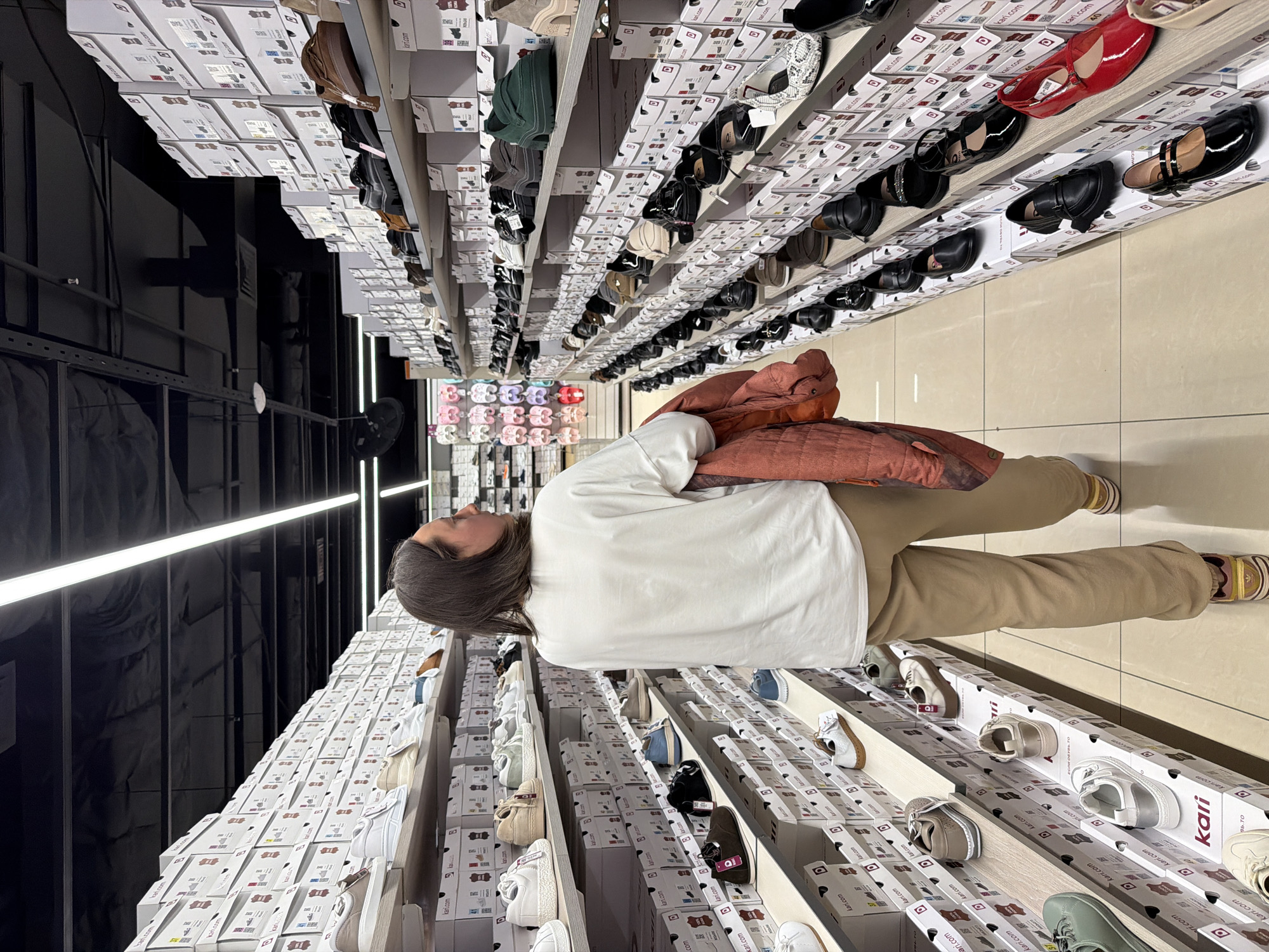 A woman is standing in a shoe store in Izhevsk, Russia, looking at the shoe displays. The store appears to have many shelves of shoes stacked in boxes, mostly white. The woman is wearing a white t-shirt and khaki pants and carrying a large orange coat. The lighting is bright and the floor is tiled.
