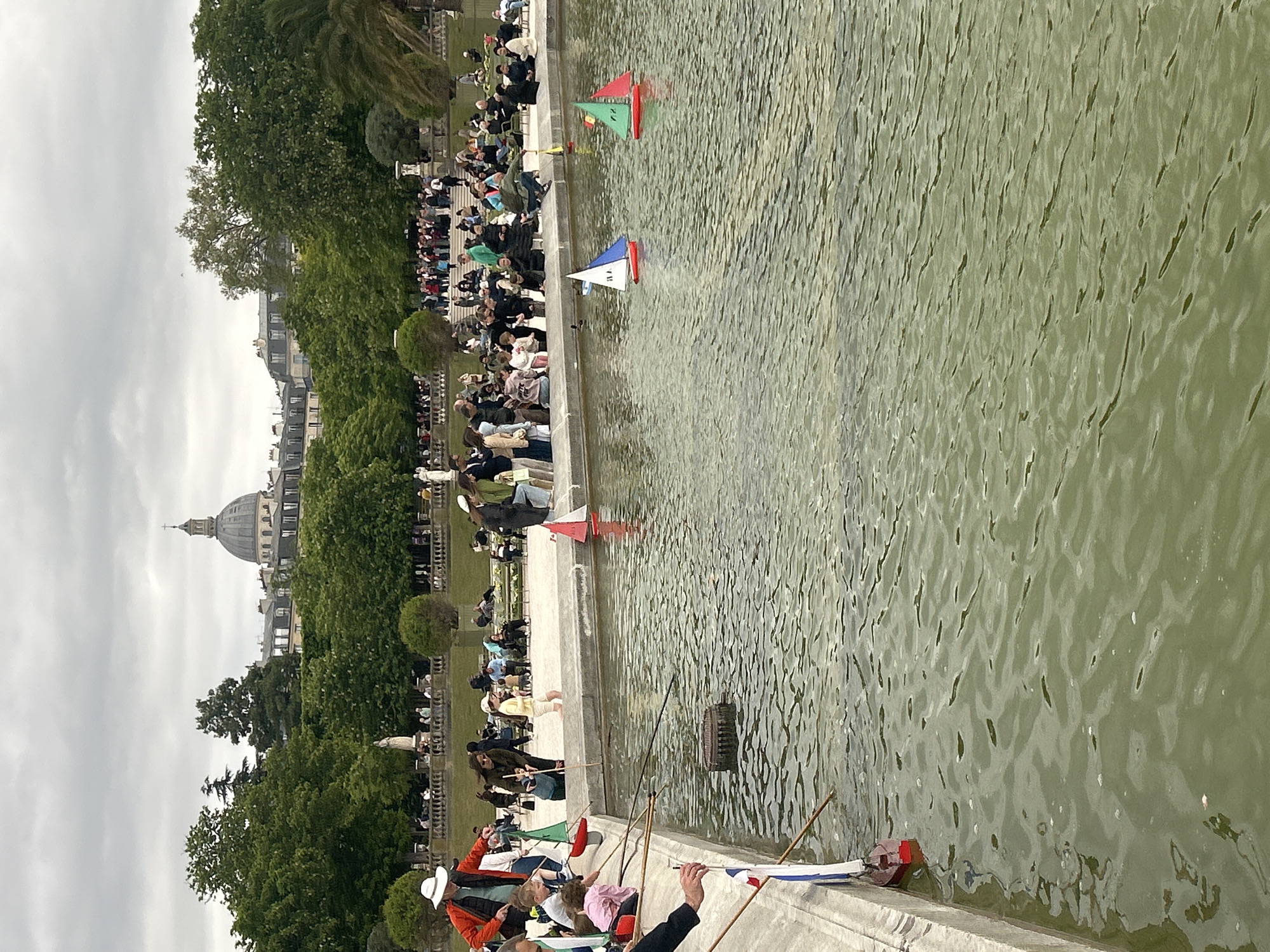 The image depicts a scene at the Jardin du Luxembourg in Paris, France. The photo is taken at ground level along the edge of a large, rectangular pond. A crowd of people is gathered along the perimeter of the pond, likely engaging in activities such as watching or participating in the operation of small model sailboats on the water. Several of these sailboats, distinguished by their colorful sails, are visible in the pond. People are launching and/or steering the boats with poles. In the background, the dome of the Pantheon is visible above the trees. The day appears overcast.