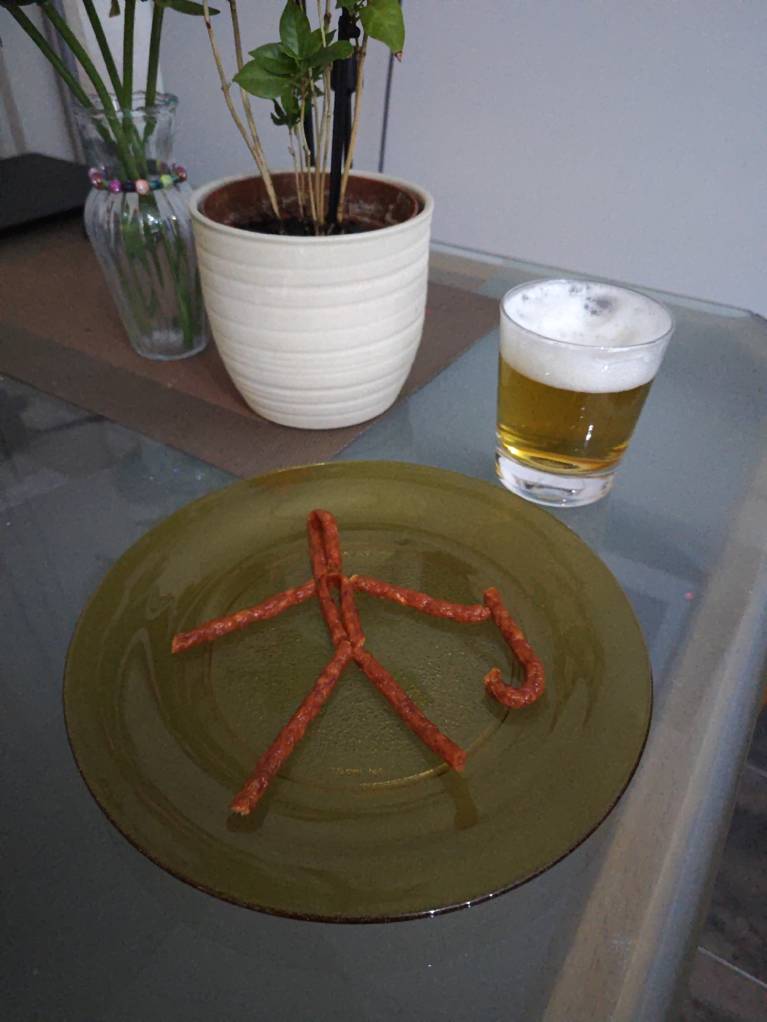 A still life image captures a green plate with a stick figure made of small, cured sausages. The sausage figure has a body, head, arms, and legs, with a curved piece for the head, two straight pieces for arms, and two angled pieces for legs. To the right of the plate, a glass of beer with a frothy head sits on a reflective surface, likely a table. In the background, a white ceramic pot contains a plant with green leaves and thin stalks. Beside the pot, a glass vase holds more plant stalks, and a beaded bracelet adorns its neck. The scene is lit by what appears to be indoor lighting, creating soft shadows. The background is a plain, light-colored wall. There is no visible text in the image.