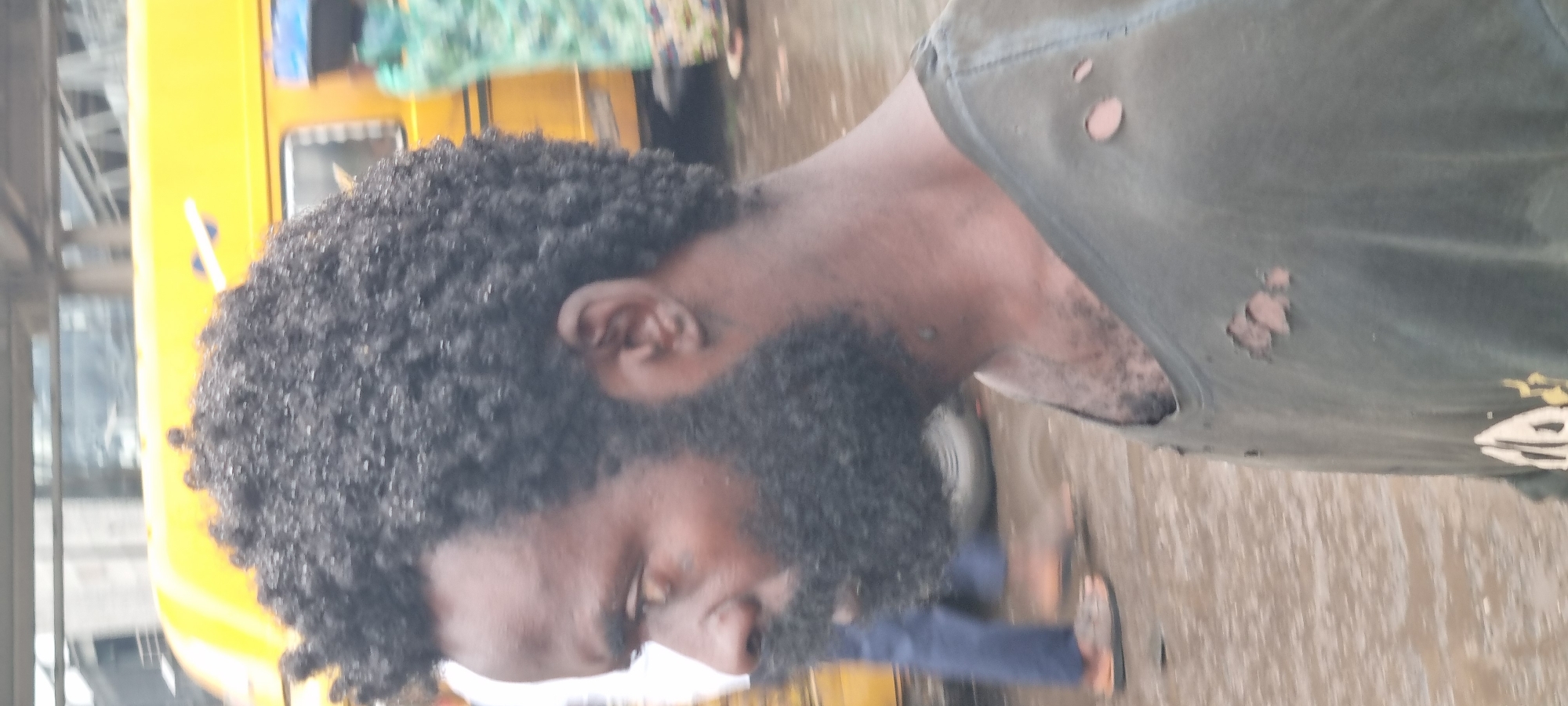 The image shows a close-up, side profile of a Black man with a dark complexion, afro-textured hair, and a full beard. He is wearing a dark, torn t-shirt. In the background, a yellow bus is visible, which is a common sight in Ikeja, Nigeria, known for its public transportation system. The ground appears wet, suggesting it may have recently rained. Other individuals are blurred in the background, indicating movement and activity in a public space. The lighting suggests it is daytime, likely overcast due to the diffused light.