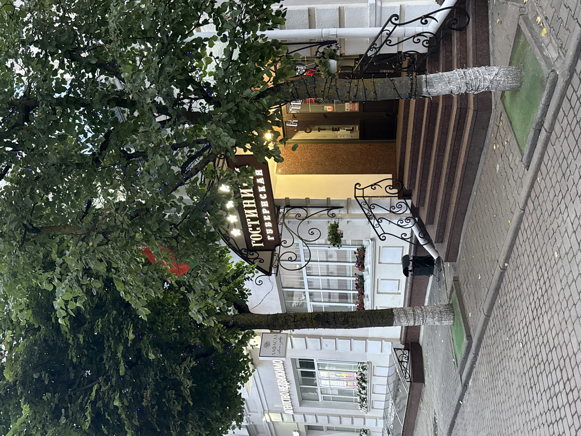 The image shows the entrance to a hotel called "Гостини Губернская" (Hotel Gubernskaya) in Mahilyow, Belarus. The hotel is situated on a cobblestone street. A tree decorated with string lights stands in front of the entrance, which features a set of stairs leading up to a double door. The building is painted white and has large windows. Another building to the left of the hotel has signage for "АГЕНСТВО НЕДВИЖИМОСТИ" (Real Estate Agency) and "ЛАФАСАД" (LAFASAD). There are flower boxes under the windows of both buildings. The overall scene depicts a street view of a commercial establishment in a city.