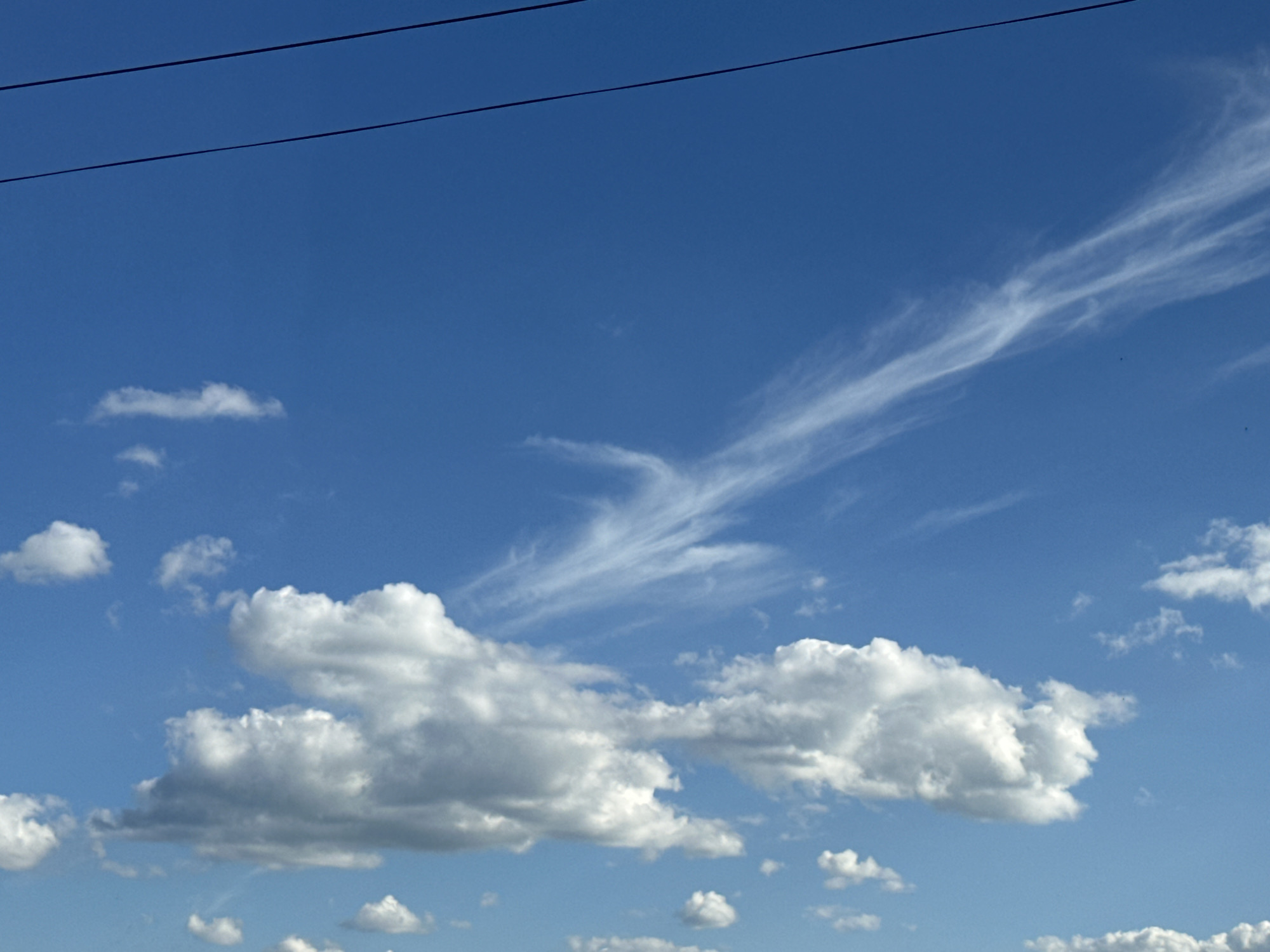 A clear blue sky with scattered clouds is visible. Two dark, thin, horizontal lines, presumably power lines, stretch across the top left portion of the image. The clouds are mostly white and fluffy, with some appearing wispy and translucent. The overall scene depicts an outdoor environment on a bright day.