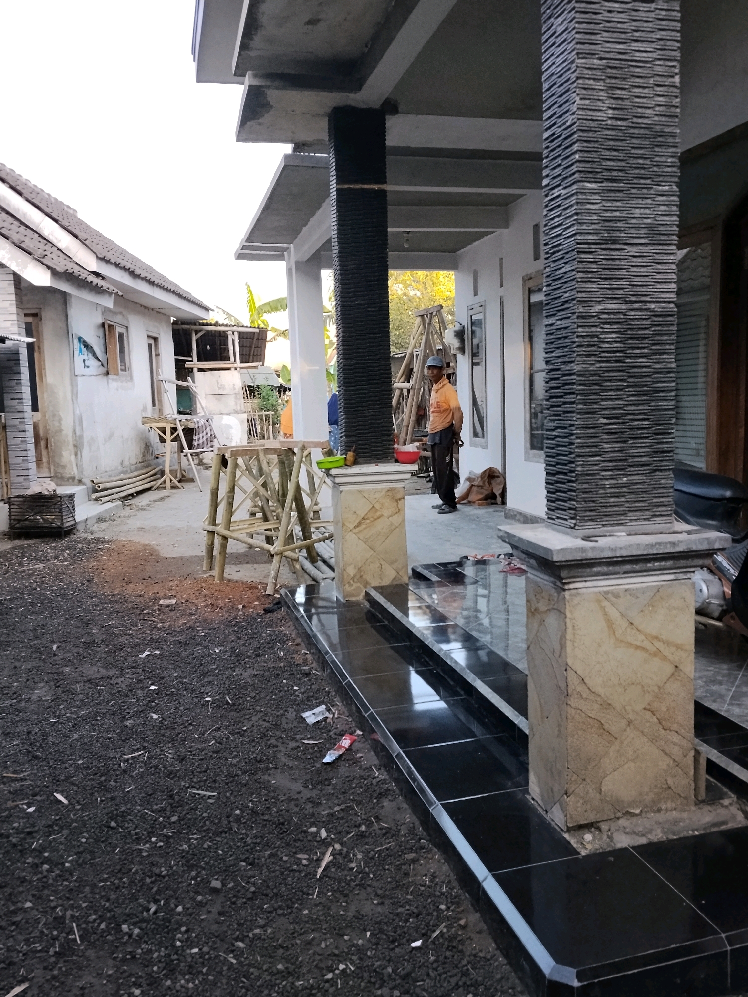 A man in an orange shirt and dark pants stands near a building under construction in Panggungan, Indonesia. He is facing left and appears to be looking at something off-camera. The scene is outdoors, with a partly constructed house visible on the left and a covered porch area in the foreground. Construction materials, including bamboo poles and scaffolding, are scattered around the area. The ground is covered with dark gravel and some debris. The lighting suggests it is either early morning or late afternoon.