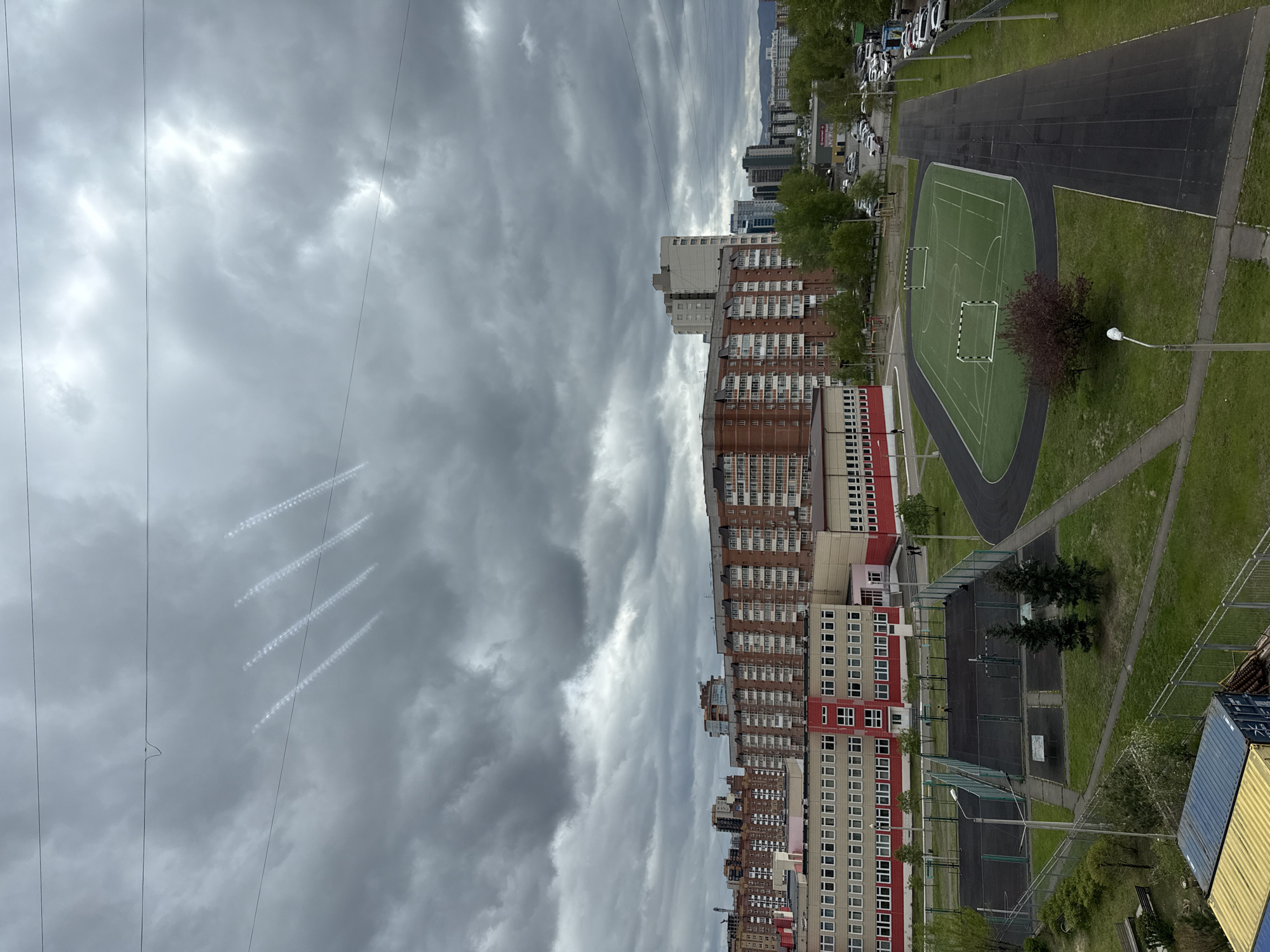 The image depicts a cityscape in Krasnoyarsk, Russia, under a cloudy sky. Streaks of condensation trails from aircraft are visible against the gray sky. The scene is viewed from a high vantage point. In the foreground, there are numerous residential buildings of varying heights and colors. A sports complex is located adjacent to the buildings, featuring a track and a green sports field with soccer goals. A row of parked cars lines the street near the complex, and other buildings are visible in the distant background.