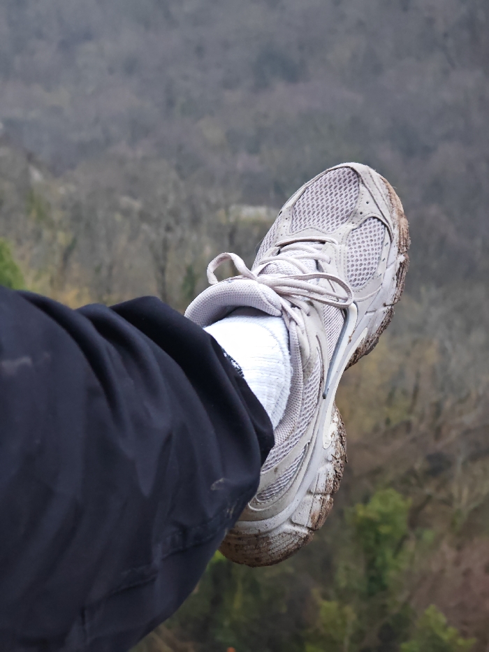 A close-up, downward-angled image captures a human leg, clad in dark, possibly black, trousers, a white sock, and a light-colored athletic shoe. The shoe, featuring a mesh upper with synthetic overlays and tied laces, has its sole and lower lateral surfaces heavily caked with dried, light-brown mud and dirt. The leg is extended forward, positioned as if over an edge or open void. The blurred background reveals an expansive panoramic view of a deciduous forest, with trees largely bare, indicative of a late autumn or winter season, observed from a significant elevation. This scene is located within the Shumen Plateau Nature Park, near the city of Shumen, Bulgaria.