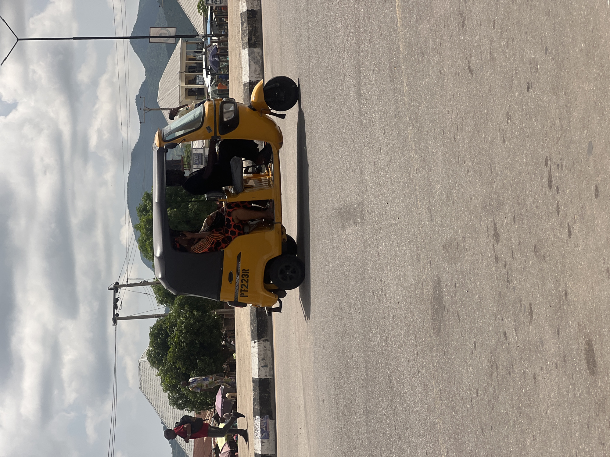 The image shows a yellow auto-rickshaw with passengers driving down a paved road in Howayi, Nigeria. The auto-rickshaw has "PT223R" printed on its side. The scene is outdoors with a cloudy sky and mountains in the background. Several people are visible near the road, some under umbrellas. The time of day appears to be daytime, judging by the lighting. The overall mood of the image seems to be casual and everyday, depicting a typical street scene in the location.
