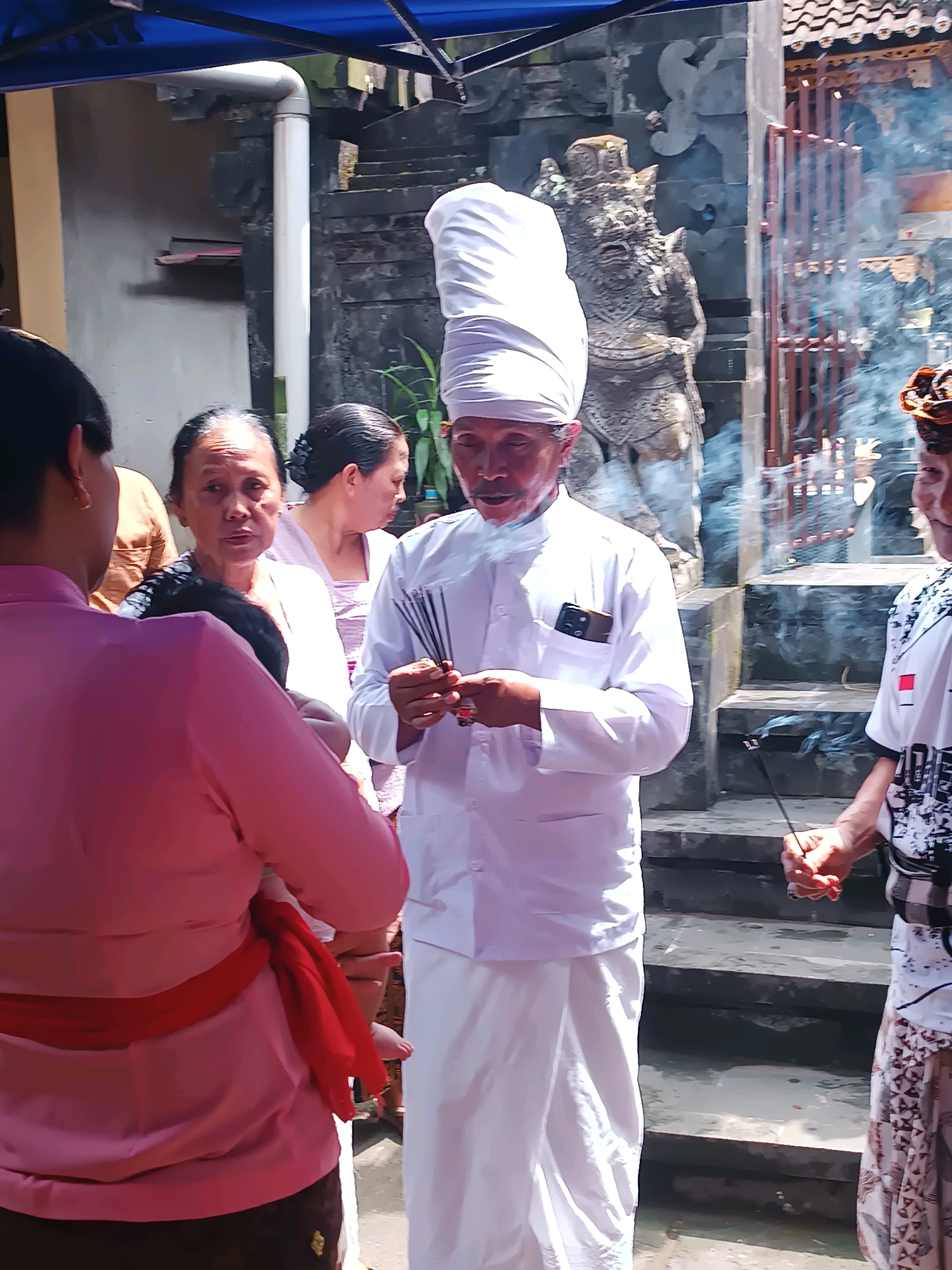 This image depicts a religious ceremony taking place outdoors, likely in Tabanan, Indonesia. In the foreground, a man in traditional white religious attire with a tall white headdress is holding several incense sticks, with smoke rising from them. He appears to be performing a ritual. To his left, a woman in a pink top and a red sash is holding a baby. Behind them, several other individuals are visible, some looking towards the man. In the background, there is a stone structure with carved figures and a staircase, characteristic of Balinese temple architecture. The overall atmosphere suggests a sacred or communal gathering.