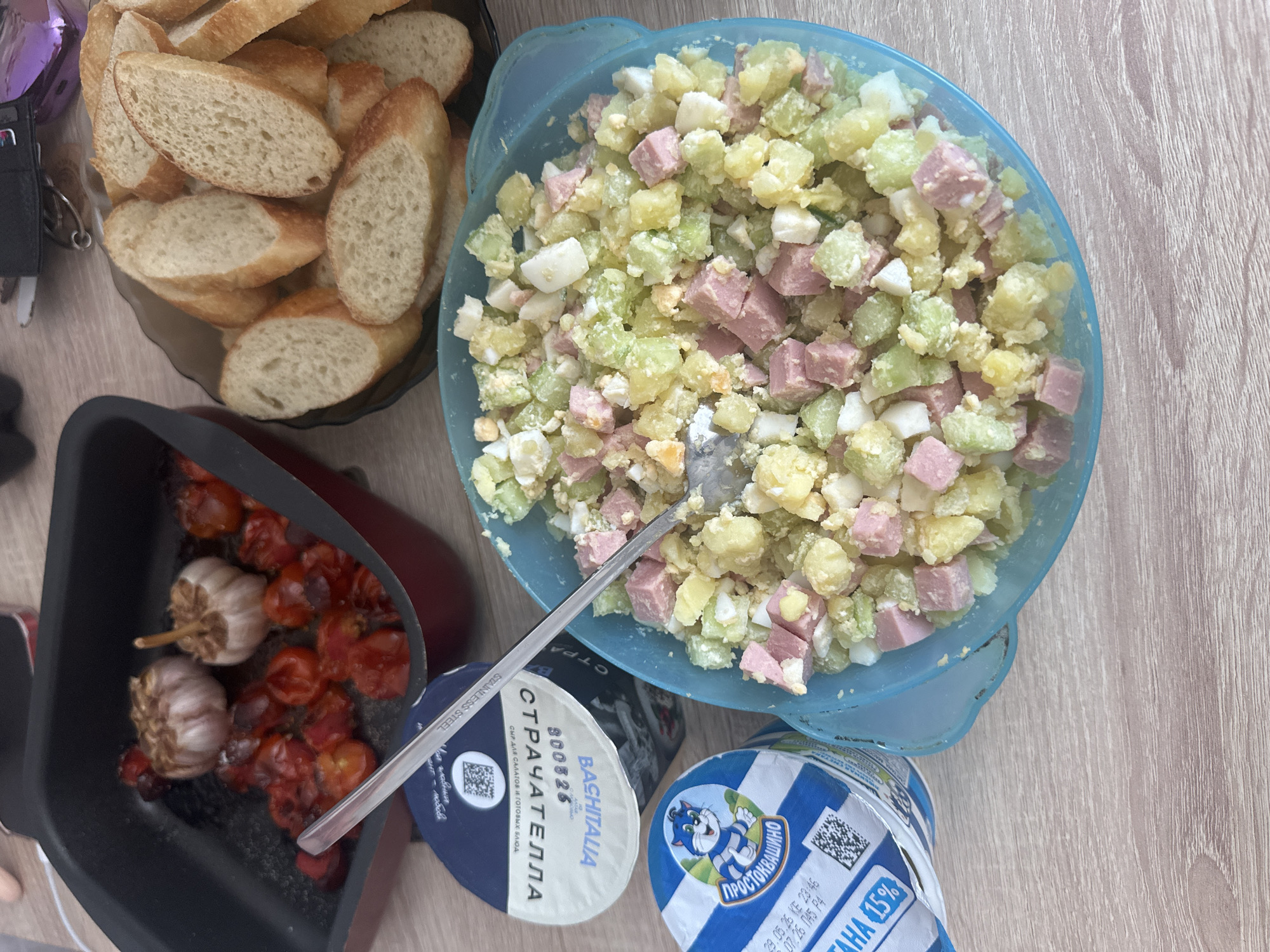 An overhead shot displays a variety of food items arranged on a light wooden table, suggesting a casual indoor meal setting during the daytime.
The central subject is a large, light blue bowl filled with a mixed salad. The salad contains uniformly diced yellowish potatoes, light green pieces (likely cucumber or celery), pinkish cubed meat (possibly ham or sausage), and white chunks of hard-boiled egg. A metal spoon rests within the salad.
To the upper left, a black plate holds multiple slices of a baguette or similar bread, cut into thick, oval pieces. In the lower-left corner, a black pan contains roasted red cherry tomatoes and two whole heads of garlic, appearing slightly caramelized.
Two plastic dairy containers are positioned at the bottom of the frame. One is white and blue, featuring a cartoon cat logo and the text "ПРОСТОКВАШИНО" (Prostokvashino), "AHA 15%", along with dates "29.05.26 KE 23:46" and "07.26 045 Р4". The other container is dark blue and white, labeled "BACHITALIA" with the text "СТРАЧАТЕЛЛА" (Stracciatella), "800826", and "СЫР ДЛЯ САЛАТОВ И ГОТОВЫХ БЛЮД" (Cheese for salads and ready meals). A metal utensil, possibly a spreader or spoon, bearing the text "STAINLESS STEEL" and partially visible Russian text "Ви главный" and "мент- любовь", rests on this container. Both dairy containers feature QR codes.
Other minor objects include a shiny purple item and a set of keys at the top left, partially visible beside the bread plate. The overall scene depicts a prepared meal with various components ready for consumption.
