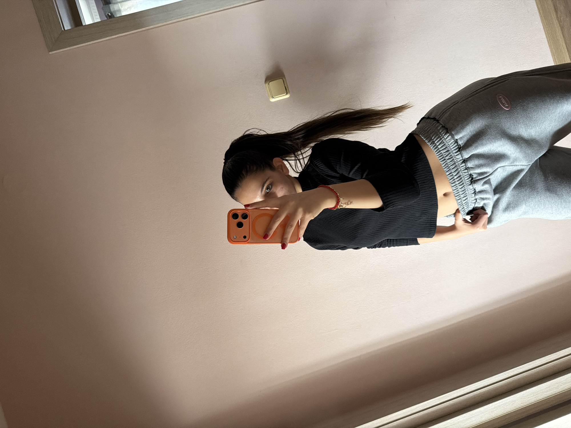A woman is standing indoors, taking a selfie in what appears to be a bedroom or similar living space. She is holding a smartphone with an orange case in her right hand. The woman has dark hair pulled back in a high ponytail and is wearing a black long-sleeved shirt and gray sweatpants. The wall behind her is a light shade of pink. There is a light switch on the wall near the top of the image. A window frame is visible at the top left, and a door frame is at the bottom right of the picture. The location is unspecified but could be in Rudartsi, Bulgaria.