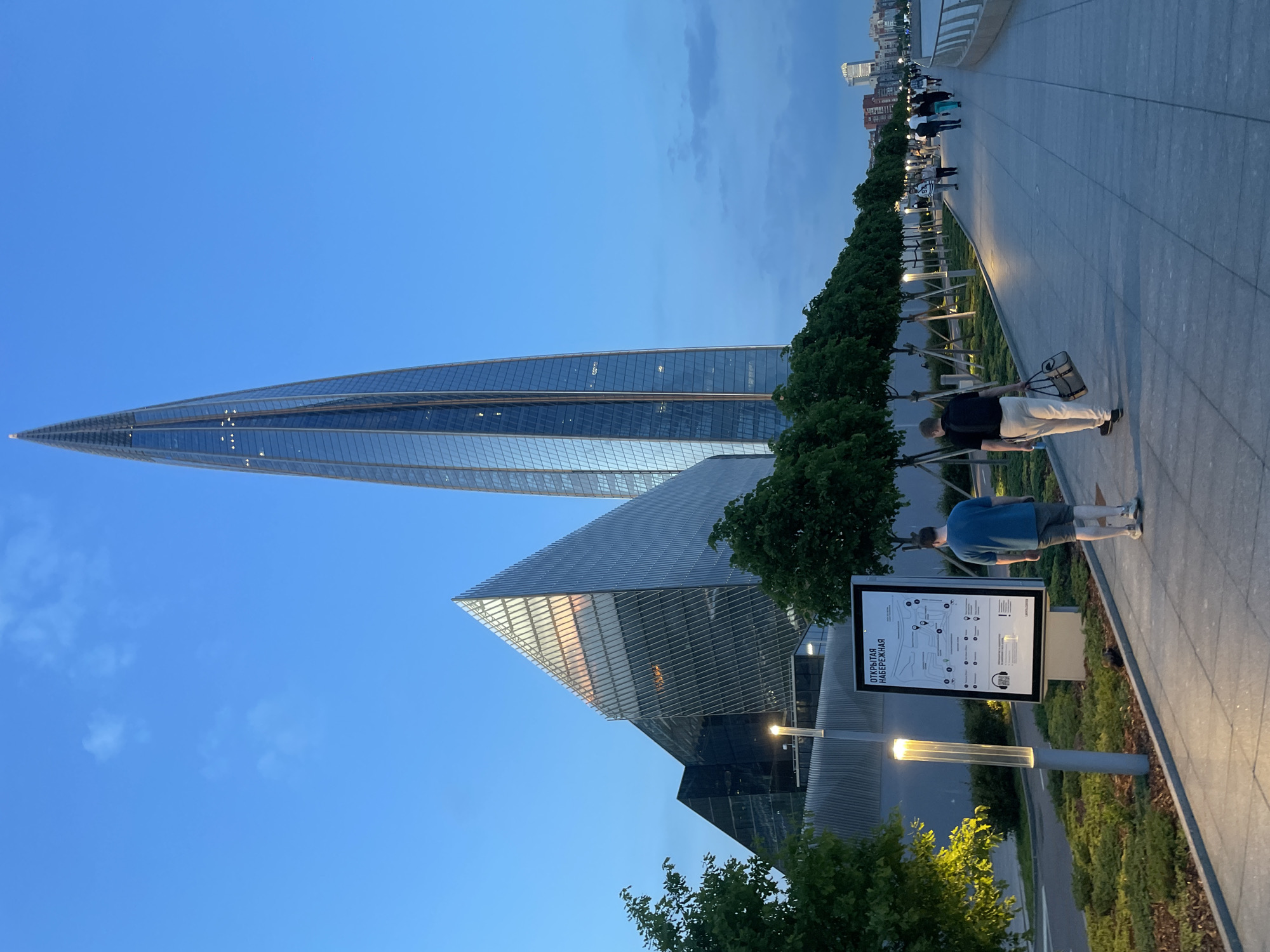 An exterior, eye-level, medium shot captures the Lakhta Center skyscraper and adjacent architectural complex in the city of Kokkorevo, Russia, at dusk. The skyscraper, a needle-like glass structure, dominates the background with its illuminated windows reflecting the twilight sky. To its left, a geometrically shaped building with angled glass panels also reflects the sky. A tree-lined walkway runs along the foreground, illuminated by modern lampposts. Two men are walking away from the camera on the walkway, one in a blue shirt and shorts, the other in a black shirt and light-colored pants carrying a bag. Several other figures are visible further down the walkway, also strolling. The ground is paved with large, grey tiles. A sign with a map and Russian text, "ОТКРЫТАЯ НАБЕРЕЖНАЯ" (Open Embankment), stands near the foreground. The sky is a deep blue with a few wispy clouds.