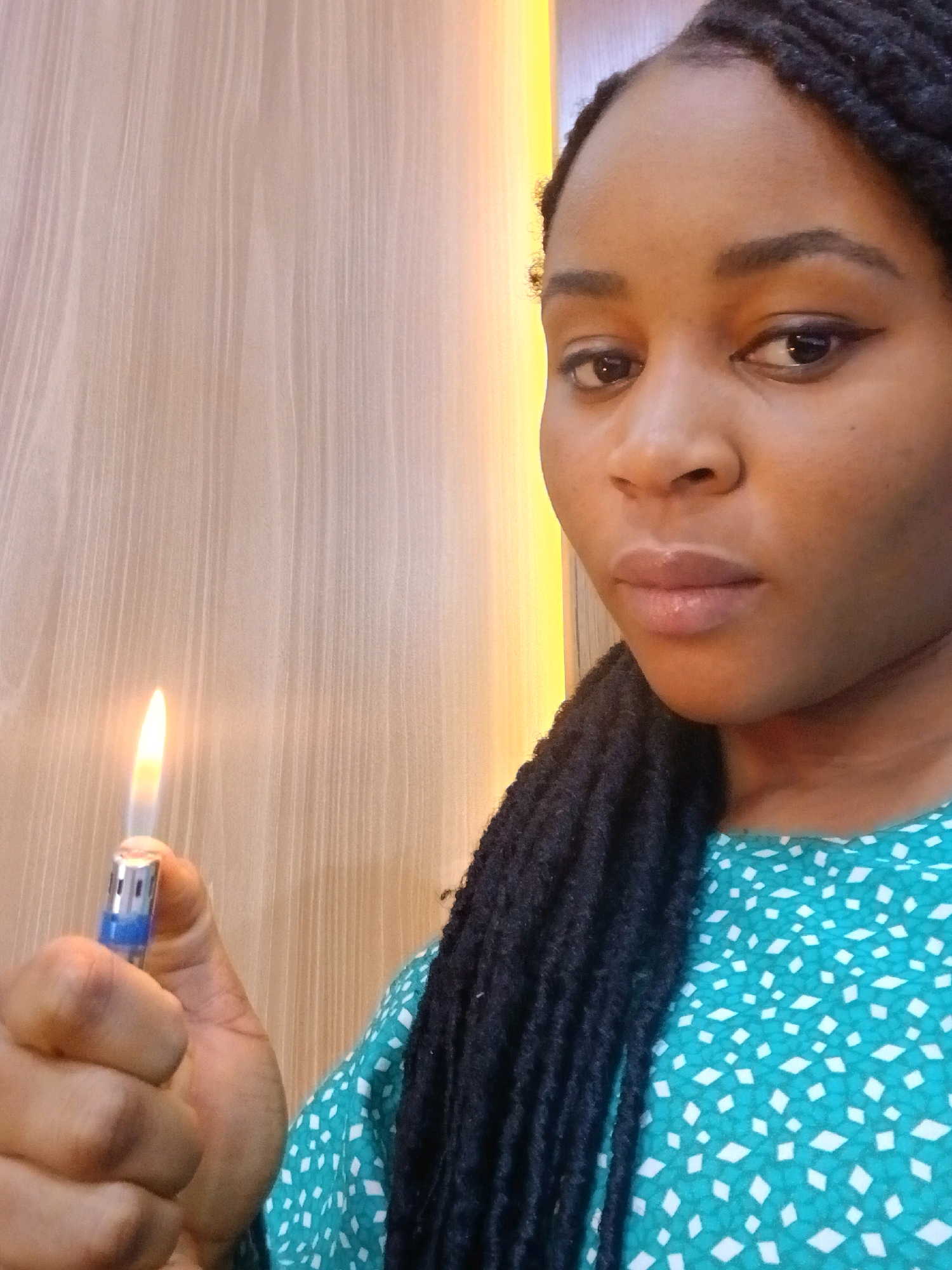 A medium close-up, selfie-style shot shows a Black person with dark braided hair holding a lit cigarette lighter in their left hand. The lighter is blue and silver and emits a bright orange flame. The person's face is partially visible on the right side of the frame, looking towards the camera. They are wearing a teal-colored shirt with a white geometric pattern. The background is a pale, wooden-paneled wall with vertical lines. The lighting suggests an indoor setting. The overall scene is focused on the person and the lighter.