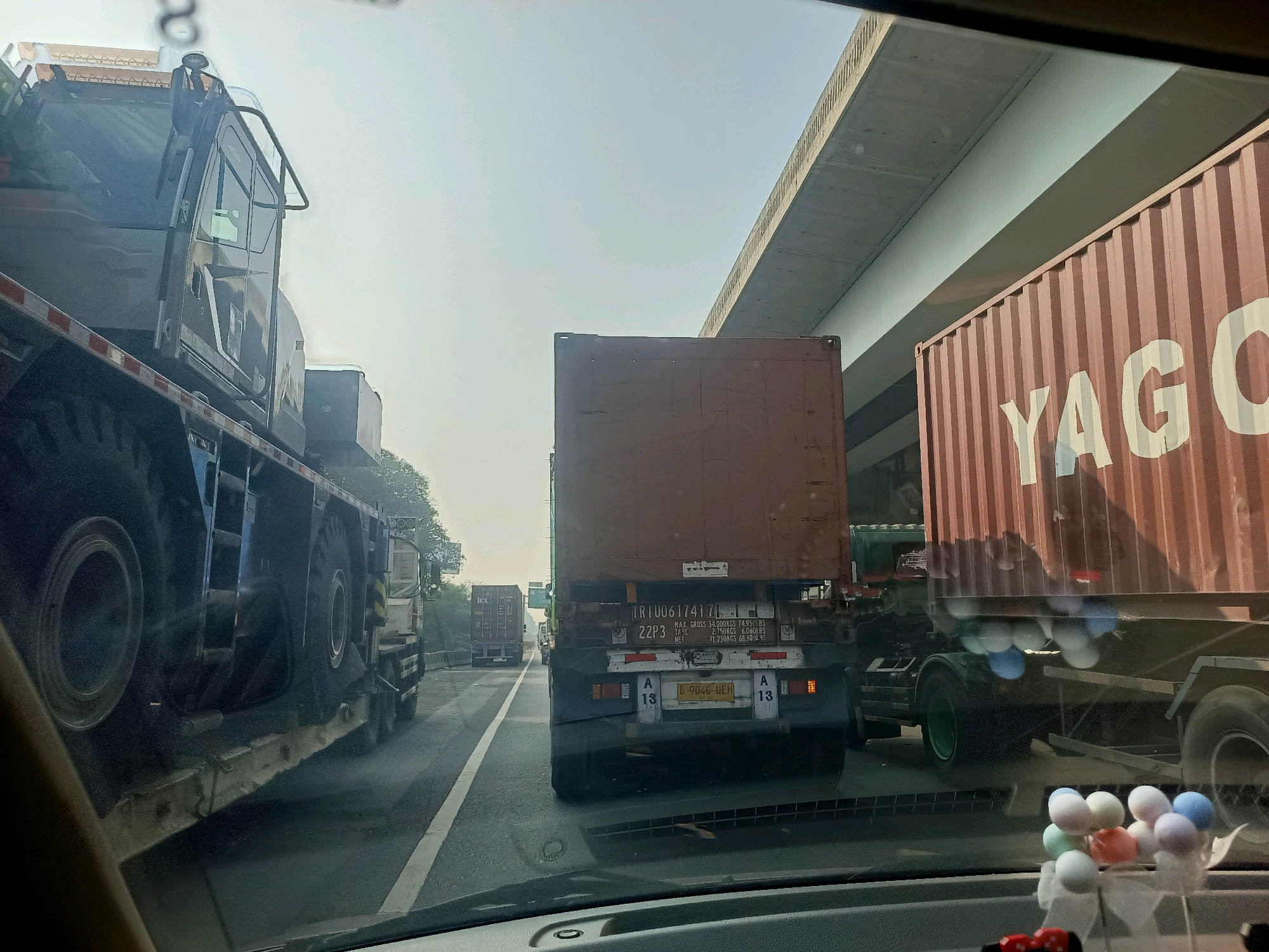 The image captures a view from inside a vehicle, likely a car, looking forward through the windshield. The scene is a busy highway, possibly in or near Cibatu, Indonesia, characterized by heavy truck traffic. The sky is overcast, suggesting a hazy or bright day.
Dominating the foreground and left side of the image is a large, dark-colored truck with an extended trailer. The truck appears to be a flatbed carrying another vehicle, possibly a piece of construction equipment, with large, rugged tires visible. The trailer has red and white striped safety markings.
In the center of the frame, a large brown shipping container is being transported on a truck. The rear of this truck is visible, showing license plates, lights, and reflective markers. The container has some labels and markings, including the text "TR1U06174171" and weight specifications.
To the right, another large shipping container, this one a reddish-brown color, is partially visible. Large white lettering on this container reads "YAGO" and parts of other letters. This container is also on a truck with visible wheels.
Several other trucks and shipping containers are visible further down the road, indicating a significant volume of freight transport. The road is paved with lane markings. In the upper portion of the image, a concrete overpass or bridge is visible, casting a shadow.
On the dashboard of the car from which the photo is taken, a cluster of colorful decorative balls is visible, suggesting a personal touch within the vehicle. The overall impression is one of a typical, congested highway environment in Indonesia, with a focus on the large-scale transportation of goods.