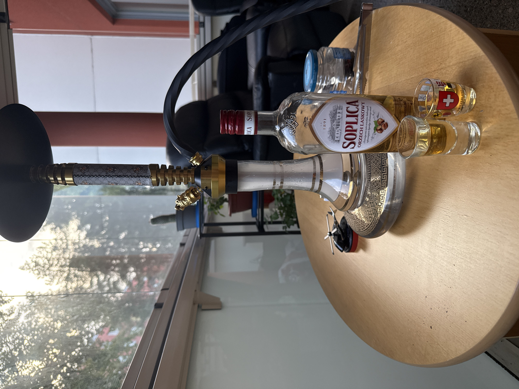 The image depicts a table on a balcony with a hookah pipe and a bottle of Soplica Hazelnut liqueur. Two shot glasses are present, one containing a yellow liquid, and the other featuring a Swiss flag design. A small toy helicopter is also visible on the table. In the background, there are trees, buildings, and a glimpse of black leather furniture. The setting appears to be a residential balcony, possibly in a city.