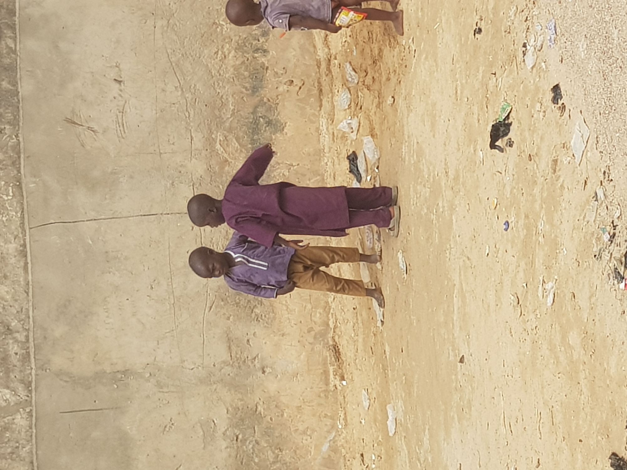 Three children are present in an outdoor setting in Dutsinma A, Nigeria. The ground is light brown and strewn with litter. Two children stand near a textured, light-colored wall. One child wears a purple outfit with khaki-colored pants. The second child wears a purple robe. The third child is in the background, and visible from the torso up, wearing a light-colored shirt and holding a food wrapper.