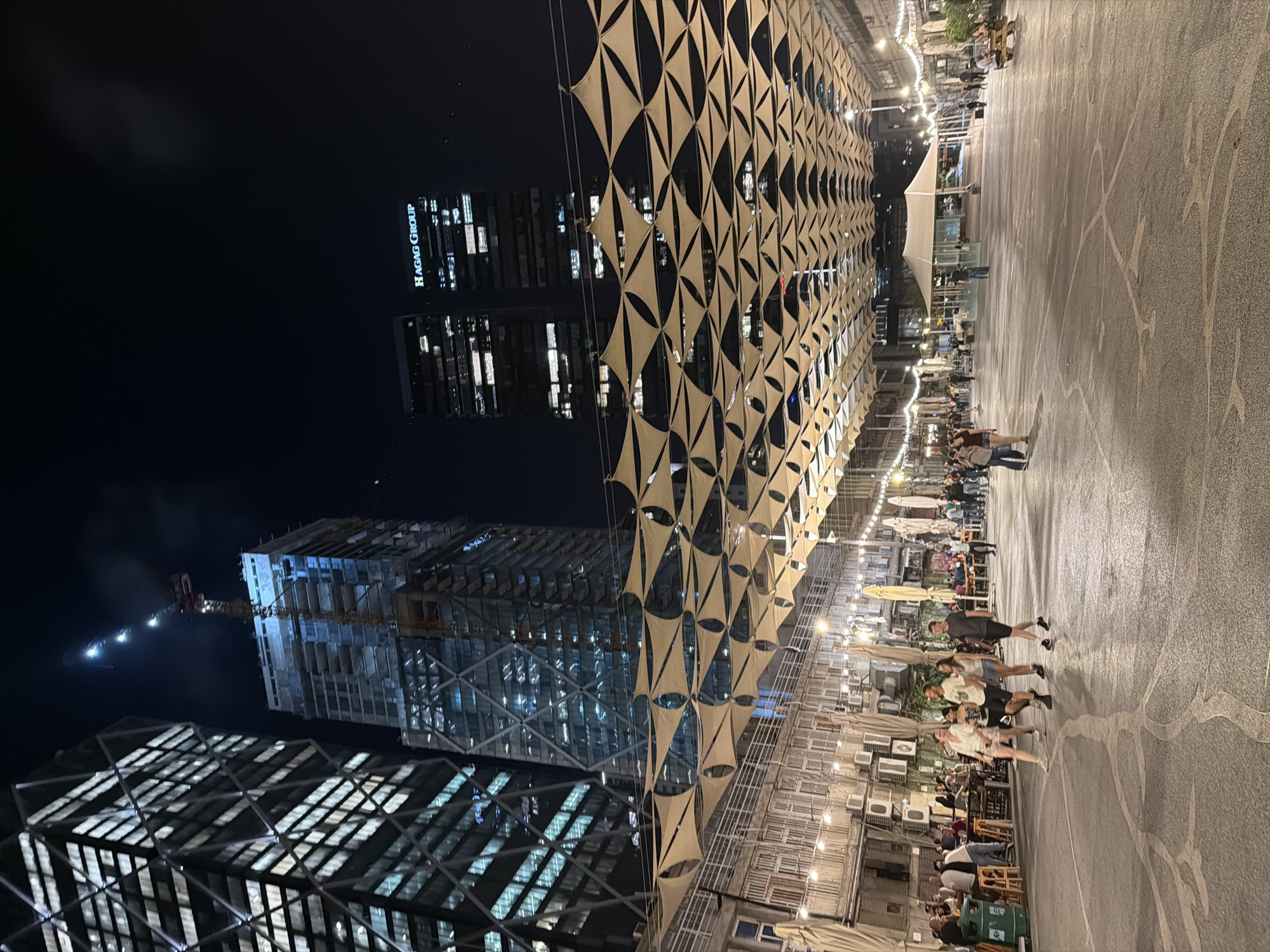 This photograph captures a nighttime scene in the Sarona complex in Tel Aviv, Israel. The foreground is a paved plaza with intricate white lines painted on the ground. A group of people is walking across the plaza. Behind them, a street lined with shops and restaurants is visible, covered by a canopy of triangular beige fabric shades. The background features modern high-rise buildings, including a skyscraper under construction with a crane at the top. Another building displays the illuminated sign "HAGAG GROUP". The scene is illuminated by artificial lights from the buildings, shops, and string lights above the canopy.
