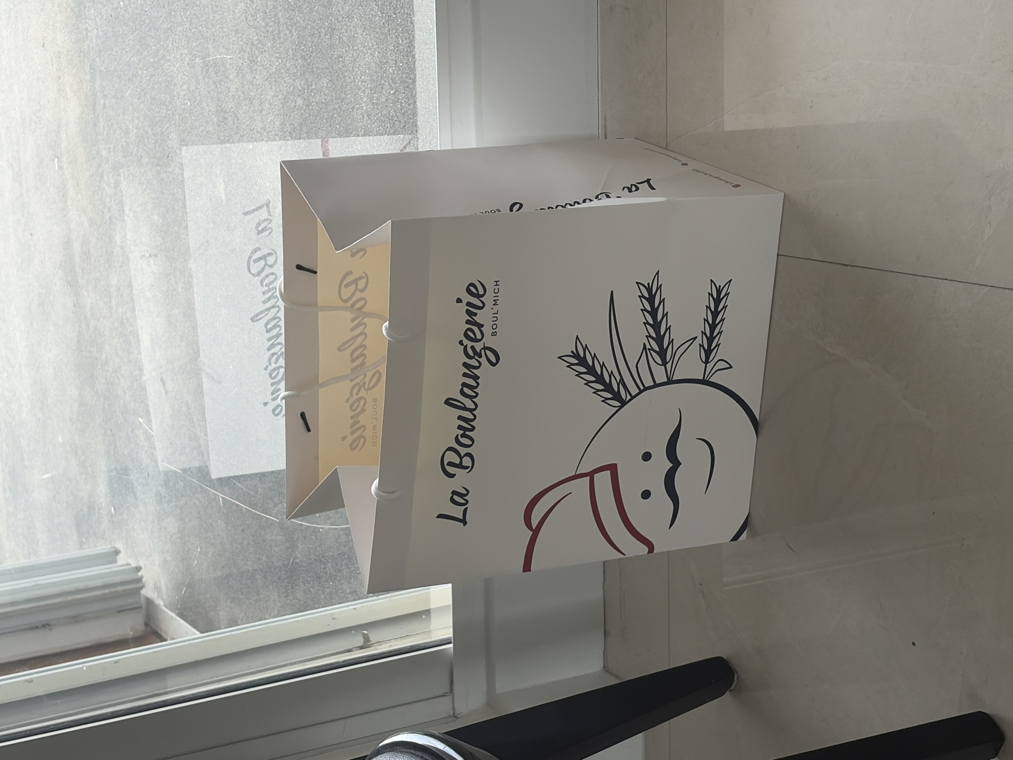 A "La Boulangerie Boul'Mich" bakery bag is partially visible in the frame. The bag is predominantly white with the bakery's name printed vertically in black on the side. Adjacent to the name is a stylized illustration that includes wheat stalks and a baker's hat. The location is near a window with a view. The bag is situated on an indeterminate surface, possibly a shelf or windowsill.