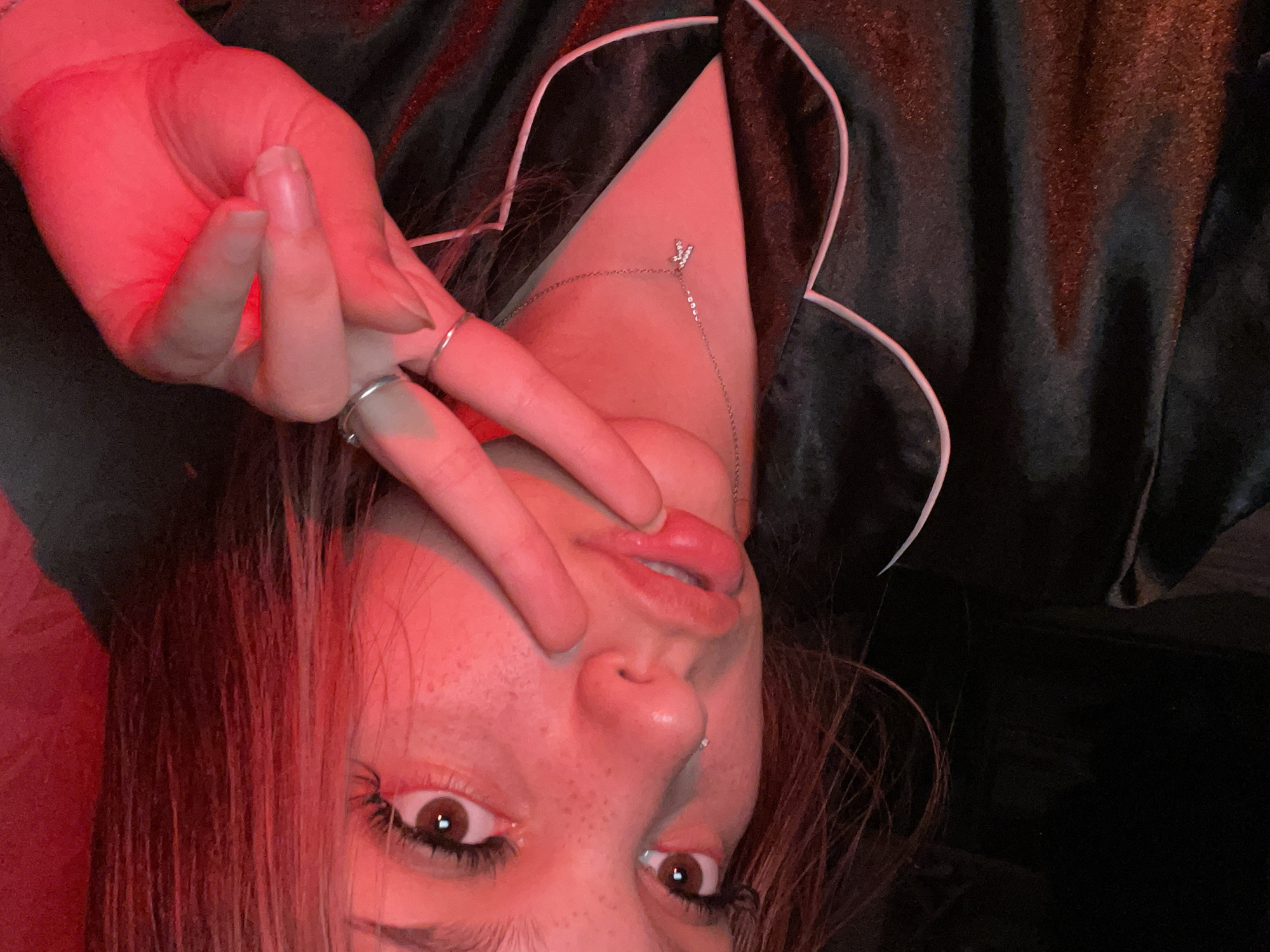 The image shows a close-up, overhead shot of a young woman with reddish-brown hair, wearing black pajamas with white trim. She has brown eyes, freckles on her cheeks and nose, and is wearing a silver necklace with a small "k" pendant. Her right hand is raised, forming a peace sign, with two silver rings on her fingers. The lighting is predominantly red, casting a warm glow on her face and pajamas. The background is dark and indistinct, with some reddish light reflecting on the fabric. The woman appears to be indoors, possibly in a bedroom, given the pajamas. No specific location in the city of Unknown, Russia can be determined from the image.