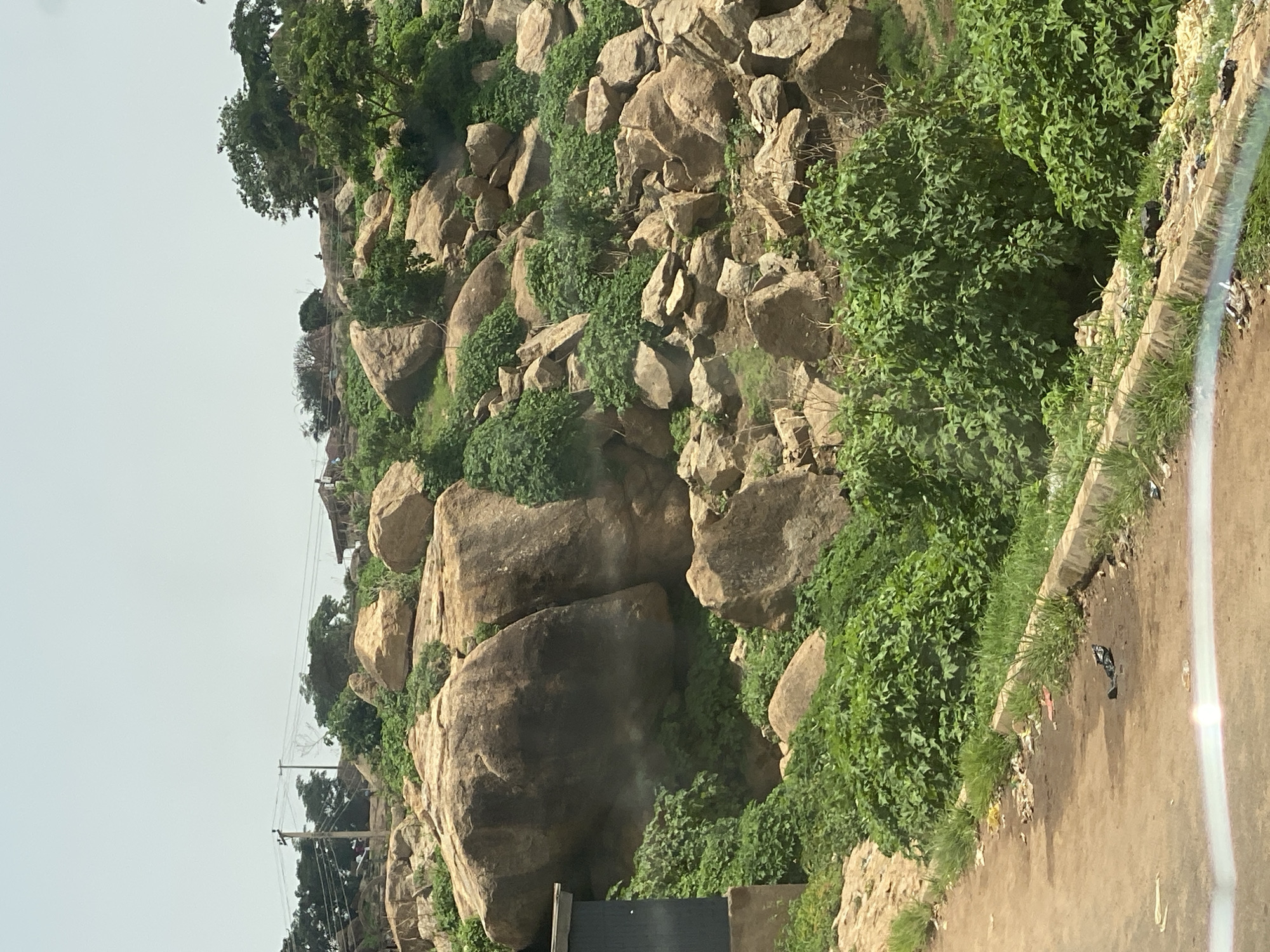 The image depicts a rocky hillside covered in lush green vegetation, with scattered large boulders. The scene appears to be outdoors and natural, likely in a rural or semi-rural area. In the foreground, a dirt road runs alongside the hillside, with a concrete channel or ditch containing some light blue liquid. The road shows signs of being a pathway, possibly for vehicles or foot traffic. Debris and trash are visible along the edge of the road and the channel, suggesting a less-maintained area.
Further up the hillside, a few scattered structures or buildings are visible amidst the rocks and trees, hinting at human habitation or development in the vicinity. Electrical poles and wires are also present, indicating the presence of infrastructure. The overall impression is of a landscape that is both natural and impacted by human activity, with a focus on the geological formations and the surrounding plant life.
The lighting suggests it is daytime, with clear weather conditions. The angle of the photograph, taken from a slightly elevated perspective looking down the road, provides a sense of depth and perspective. The colors are dominated by the earthy tones of the rocks and soil, contrasted with the vibrant green of the foliage. The presence of trash points to potential environmental concerns in the area. Without further context, it is difficult to pinpoint a specific activity or event, but the scene suggests a location that is regularly traversed or inhabited.