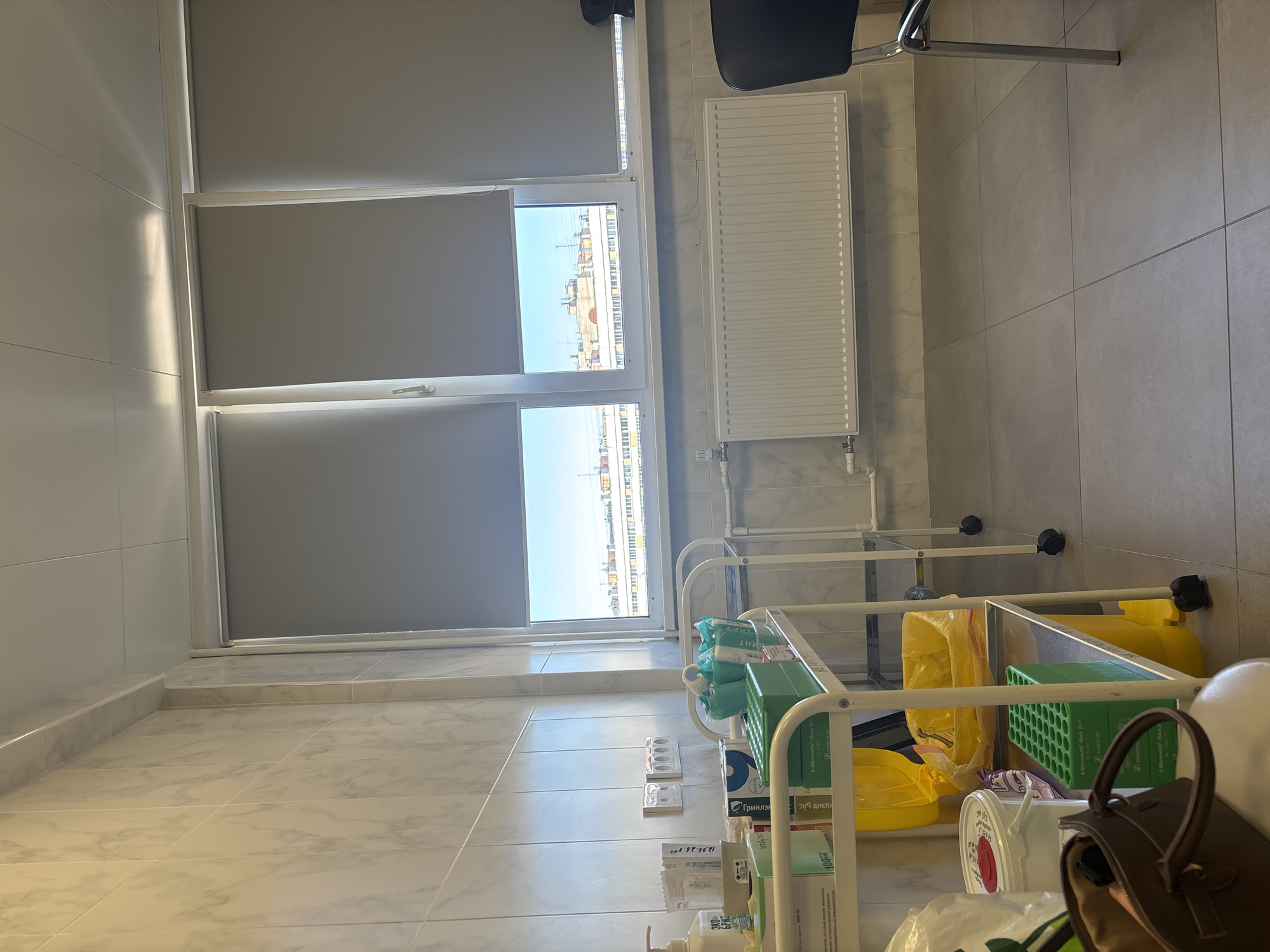 The image captures the interior of a room, likely a medical office or clinic, located in Saint Petersburg, Russia. The room features light-colored, square-tiled walls and flooring. A window with a white frame is partially covered by gray roller blinds. Outside the window, a cityscape of Saint Petersburg is visible.
A white radiator hangs on the wall near the window, and a black chair is positioned near the radiator. A white metal cart with multiple tiers is located near the window and contains various medical supplies and disposal containers, including yellow biohazard bags, green boxes, and white containers. Medical supply packaging labeled in Russian is also visible. A brown handbag is situated near the cart.
No people are visible in the image.