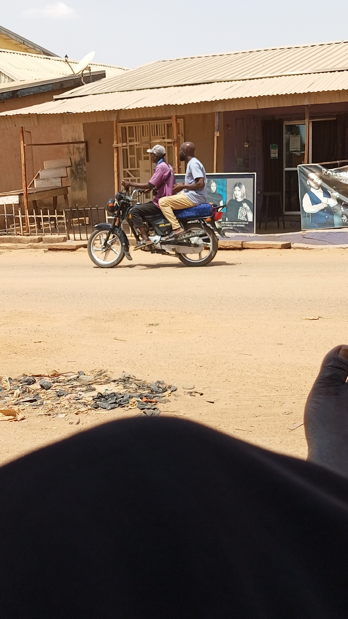 Sabon Tasha, Nigeria: Two men ride a black motorcycle on a dirt road. The driver wears a purple shirt and a light-colored cap. The passenger wears a light blue shirt and tan pants. Buildings with corrugated metal roofs line the street. Debris is scattered on the ground near the foreground. A person's leg and foot are visible in the foreground, partially obscuring the scene.