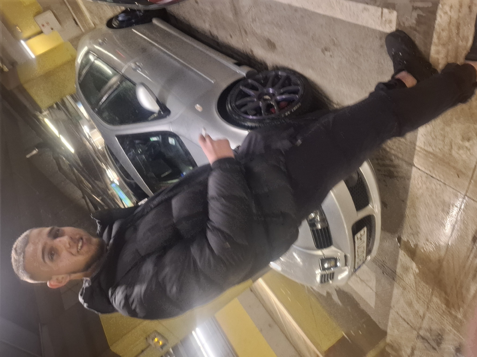A man stands next to a silver car in a garage-like setting. The man is wearing black pants, a black puffer jacket, and black shoes. He is holding a lit cigarette in his left hand. The car is parked in a garage, with yellow walls. The image's perspective is slightly rotated. I can not identify the specific location within Sofia.