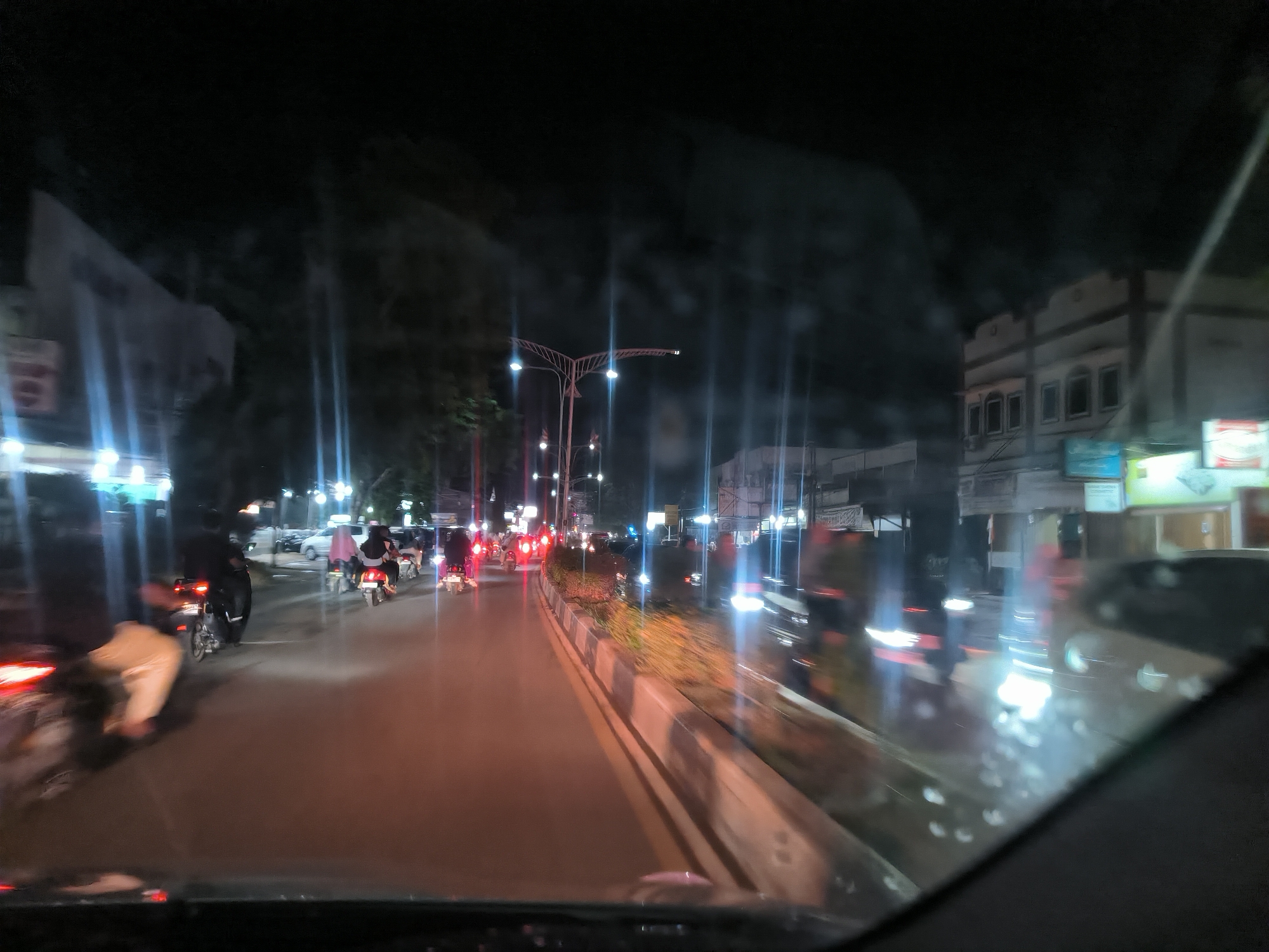 The image shows a night scene of a street in Langsa, Indonesia. It is taken from inside a vehicle, looking out through the windshield. The road is illuminated by streetlights, and there are motorcycles and cars traveling along the street. Buildings line the side of the road. A median with greenery divides the road. The scene appears to be a typical urban environment at night.