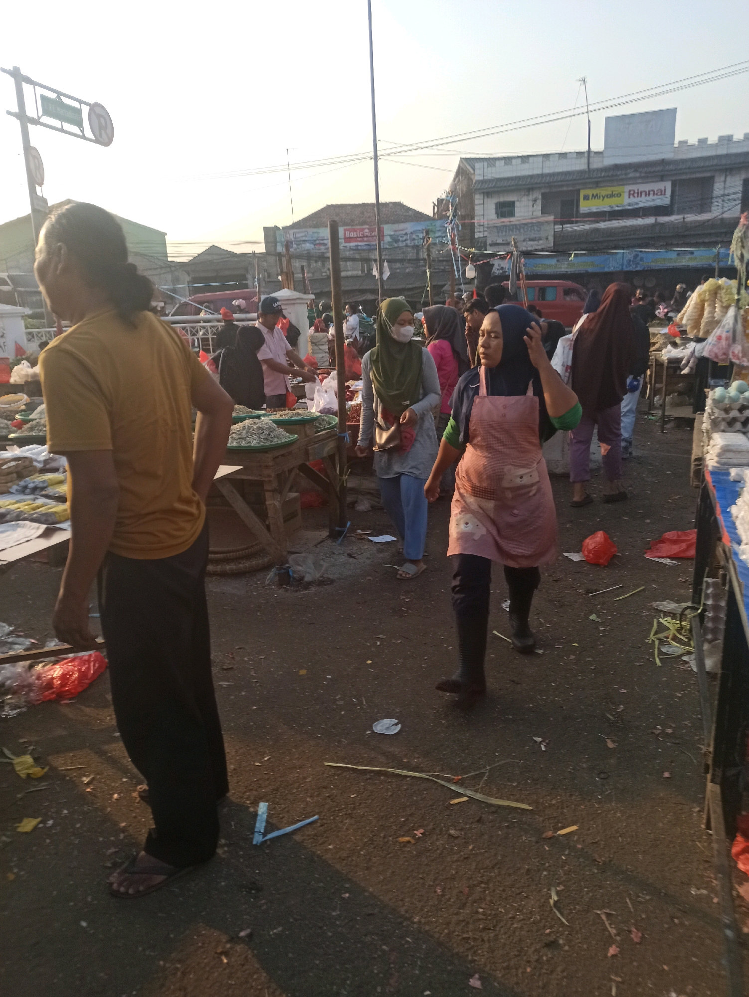 The image depicts an outdoor market scene in Cikarang, Indonesia, likely during daylight hours, possibly early morning or late afternoon given the lighting. A man with dark hair pulled back and wearing a mustard-yellow t-shirt and dark pants stands on the left, facing away from the camera with his right hand on his hip. A woman in the foreground, wearing a dark hijab, a green t-shirt, and a pink apron adorned with cartoon characters, is walking towards the right, with her right hand touching her head. She is also wearing dark pants and black boots. Behind her, other individuals are visible, including a woman in a green hijab and light blue pants, and others in the background engaged in market activities. Several stalls with merchandise, possibly food items like dried fish and eggs, are set up. The ground is paved and appears somewhat dirty, with scattered debris, including plastic bags, leaves, and discarded items. In the background, buildings with signage, including one that reads "Miyako Rinnai," suggest a commercial area. A traffic sign with a red circle and a diagonal line, indicating a prohibition, is visible on a pole. The overall impression is of a bustling local market.