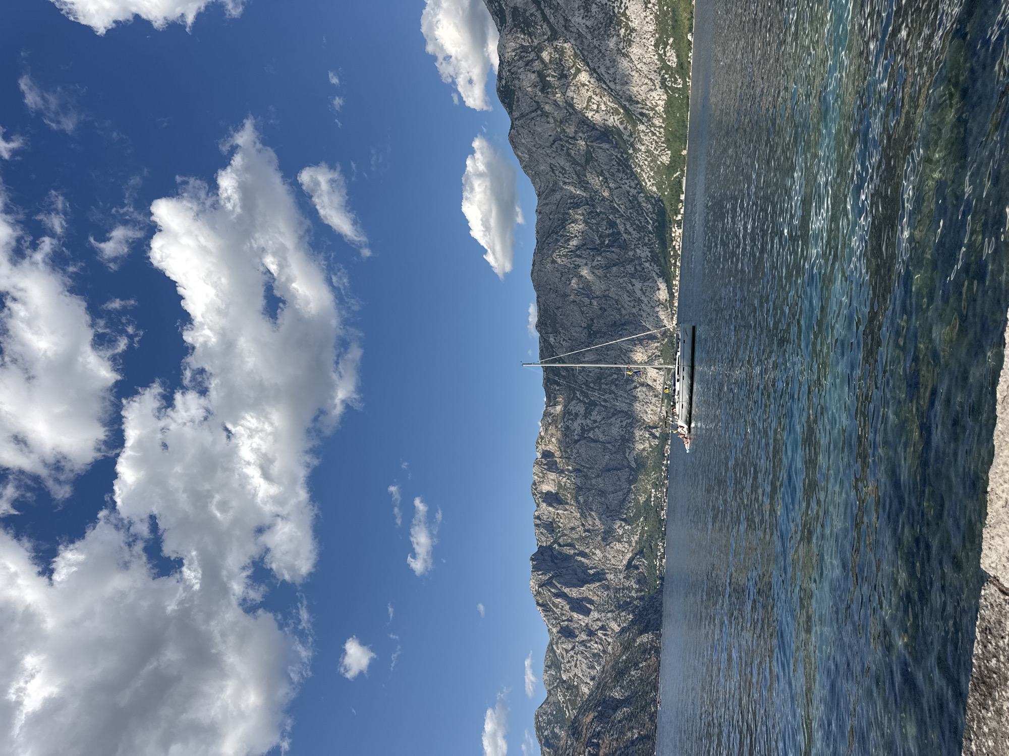 The image depicts a serene body of water with a sailboat anchored in the middle. The water is clear and reflects the blue sky and fluffy white clouds above. In the background, a majestic mountain range rises, its rugged peaks covered with sparse vegetation. A small town is visible nestled at the foot of the mountains. The foreground shows a stone embankment, suggesting a waterfront view. This scene is located in the town of Prčanj, Montenegro, a coastal settlement known for its historical architecture and stunning natural beauty.