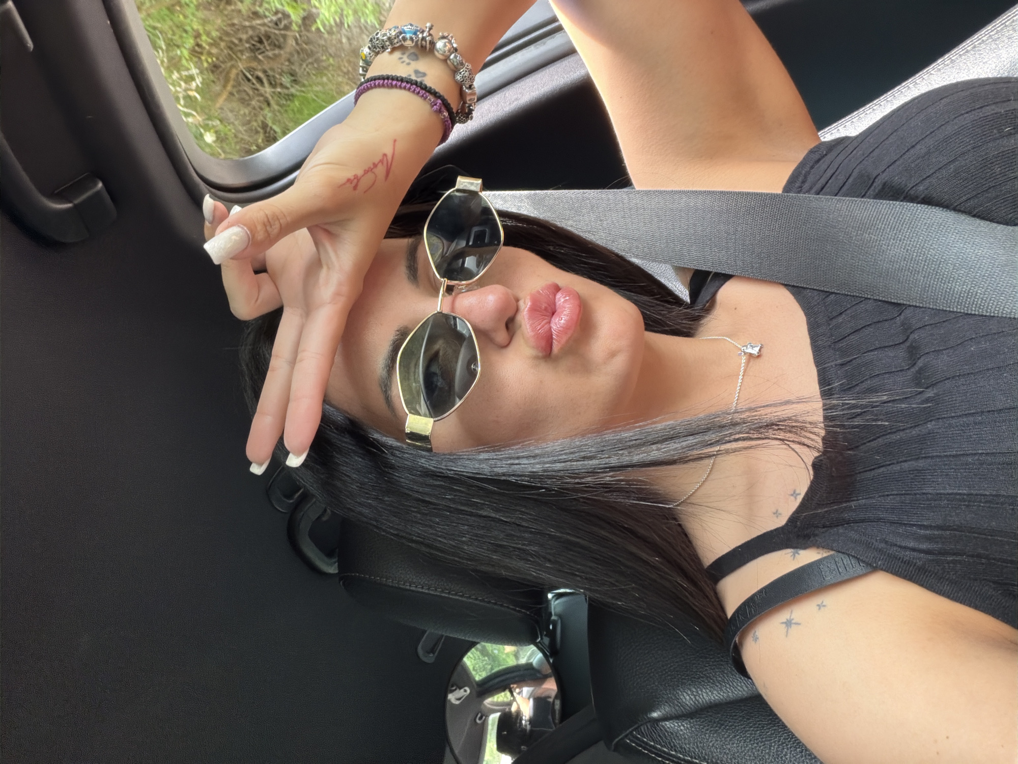 A woman wearing sunglasses and a seatbelt is reclining in the driver's seat of a car. She has dark hair, light skin, and is wearing a black top. Her left hand is raised to her forehead, with visible tattoos on her wrist and forearm. Bracelets are visible on her left wrist. The interior of the car is black. Through the window, green foliage can be seen. The location in the city of Gurgulyat, Bulgaria cannot be confirmed from the image.