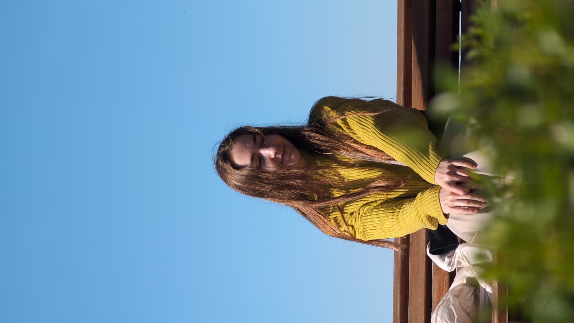 The image shows a young woman sitting on a wooden bench. She has long brown hair and is wearing a yellow sweater. She is sitting with her hands resting on her legs. Behind her, there is a bright blue sky. To her right, there are green bushes. The location is possibly a park in Batumi, Georgia.