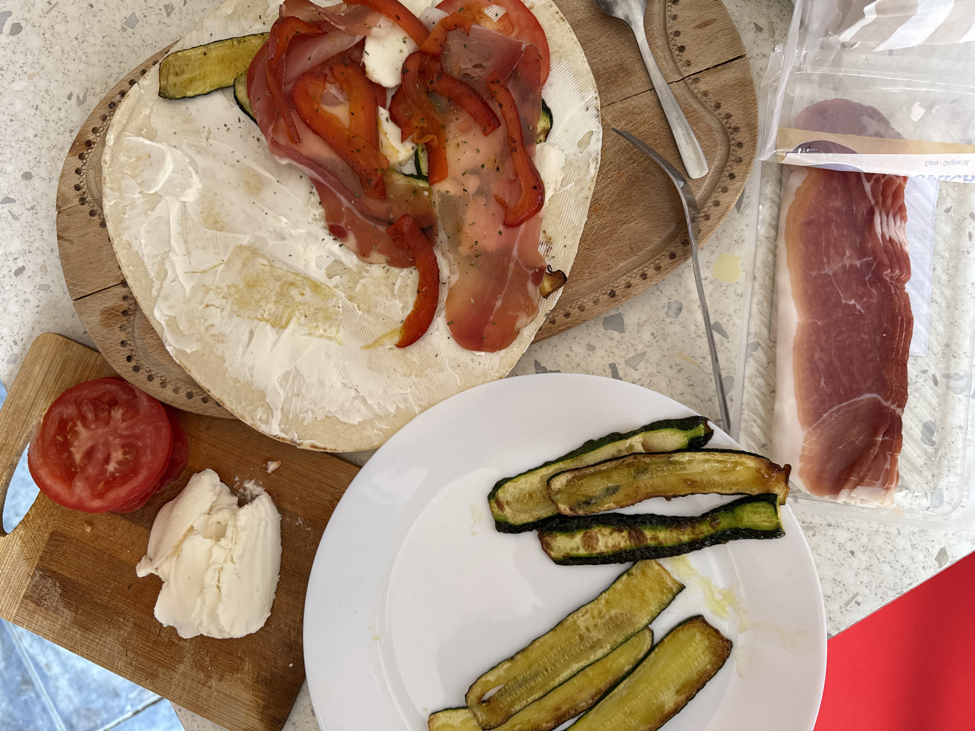 The image presents a culinary arrangement, seemingly in preparation for a meal. On a wooden cutting board, a large, round flatbread is spread with a creamy white substance, likely cheese. Atop this, strips of prosciutto, red bell peppers, and zucchini are arranged. Adjacent to the board is a white plate containing more grilled zucchini strips. A separate wooden board holds sliced tomatoes and a mass of soft white cheese, possibly mozzarella. A package of prosciutto is also visible, partially opened. Silverware is present. The overall scene suggests a relaxed, casual dining setup. Without additional context, it is impossible to confirm the location is Milan, Italy.